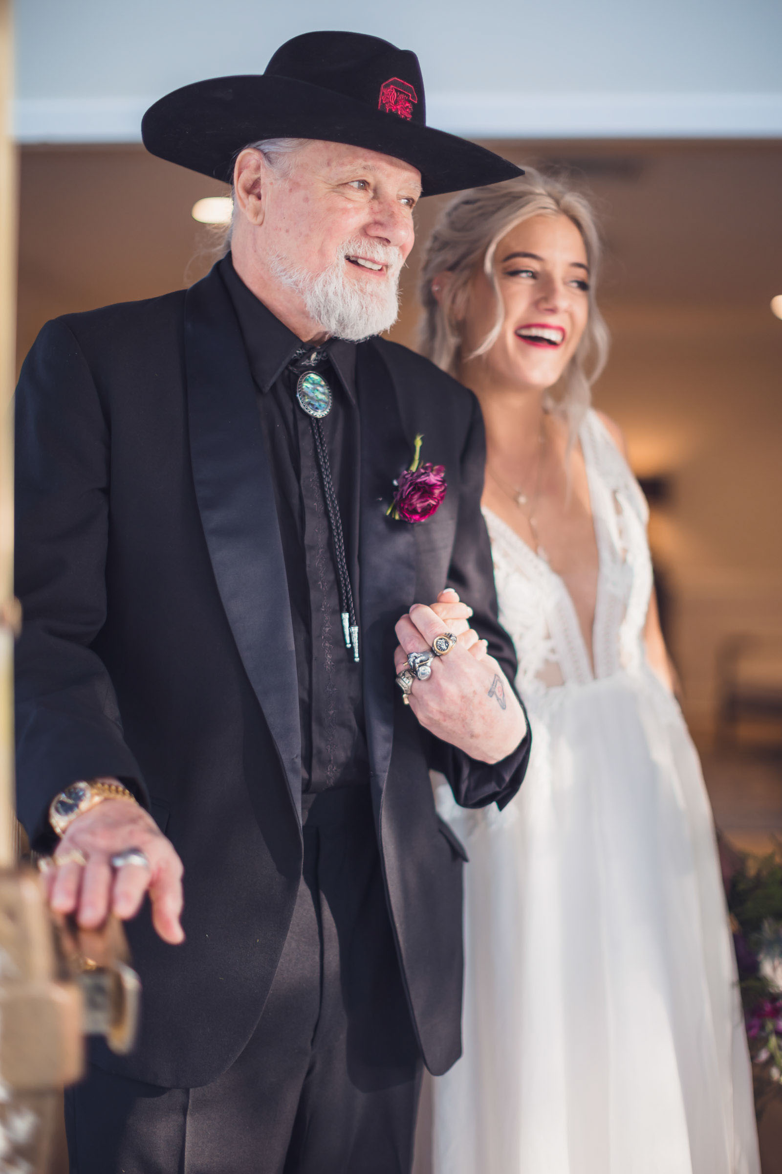 Bride smiling and walking arm in arm with her father in black suit and cowboy hat during wedding ceremony entrance at Alhambra Hall Charleston. He is Wearing a Bolo Tie.