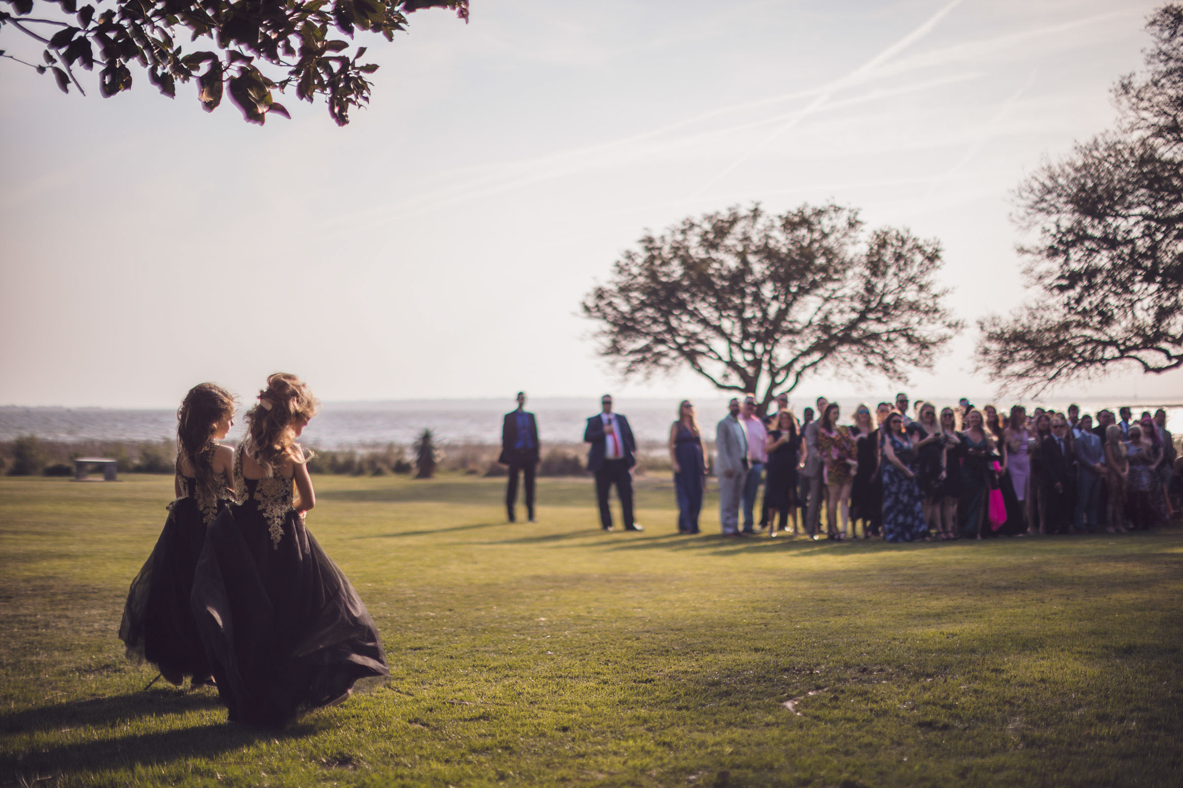 Flower girls in black dresses walking across lawn toward waterfront ceremony with guests waiting at Alhambra Hall Mount Pleasant
