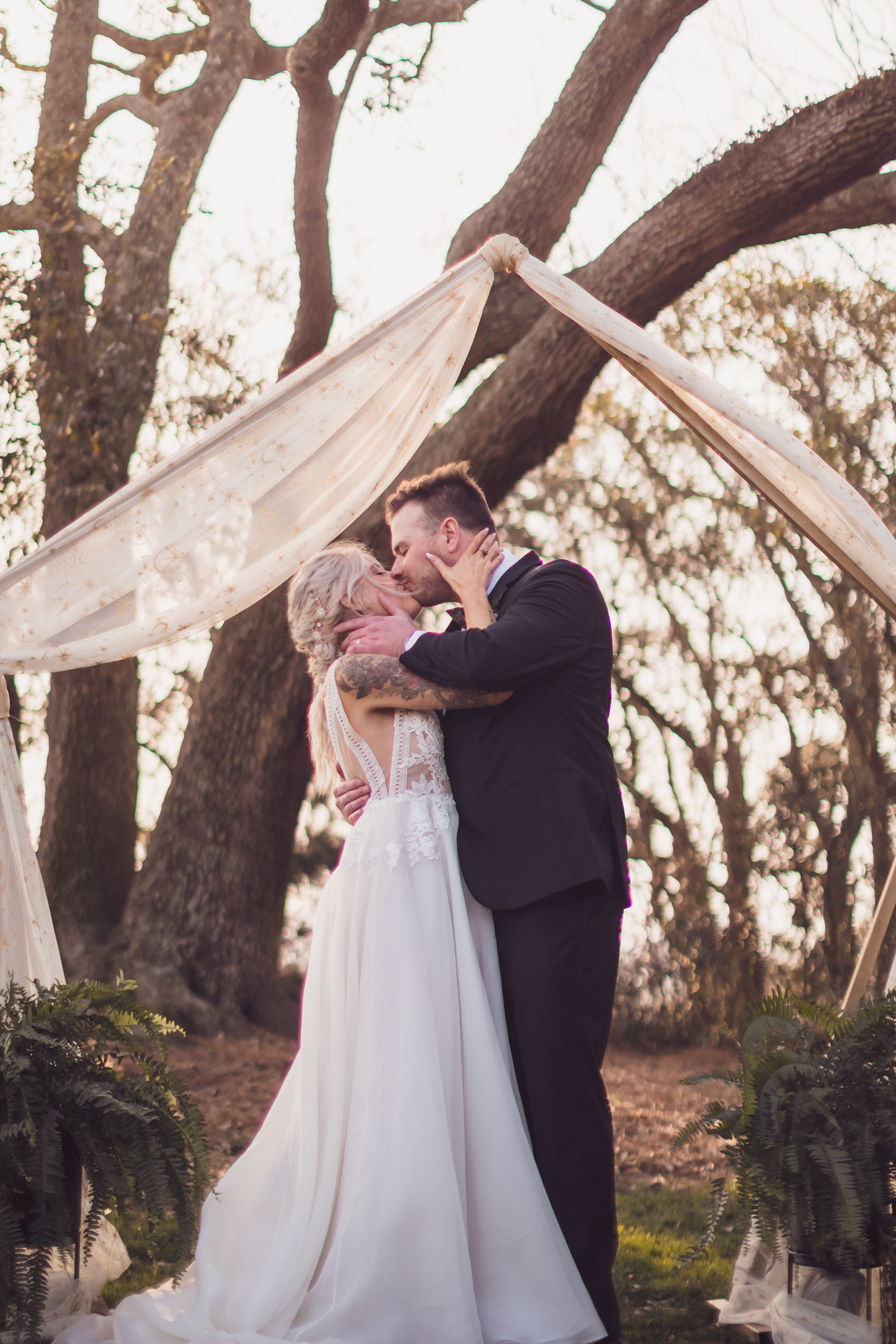 Bride and groom kissing under fabric-draped ceremony arch with oak trees in background at Alhambra Hall Charleston wedding