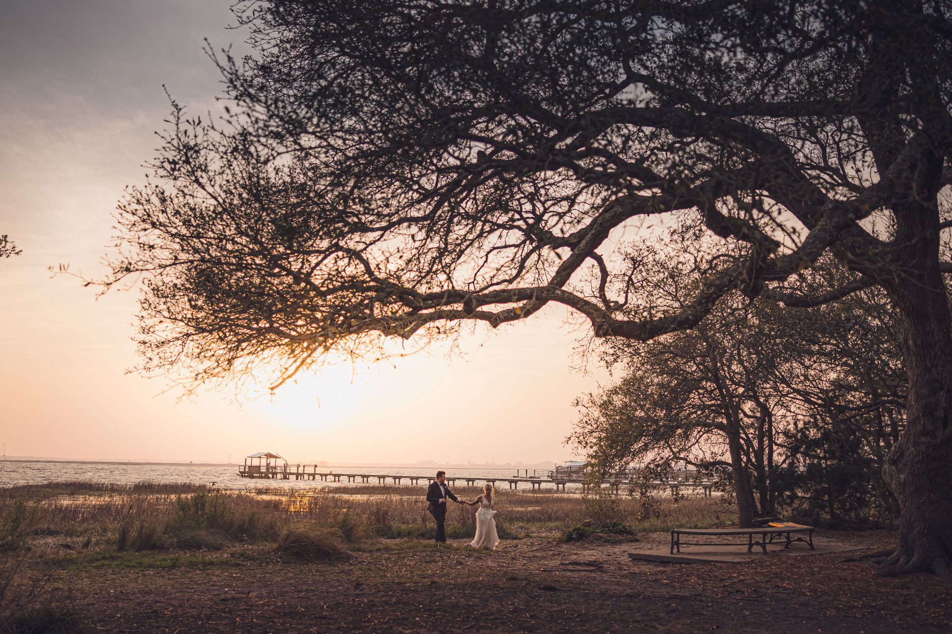 Bride and groom walking hand in hand along waterfront under large oak tree at sunset near Alhambra Hall Mount Pleasant Charleston