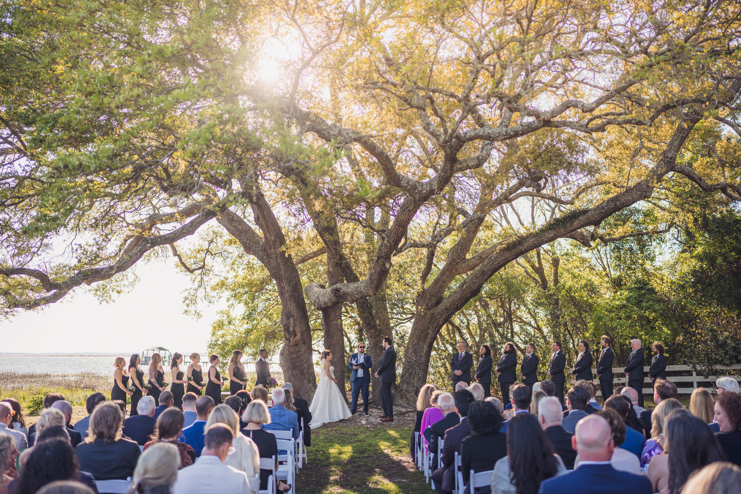 Outdoor wedding ceremony under sprawling oak tree with guests seated and waterfront view at Alhambra Hall