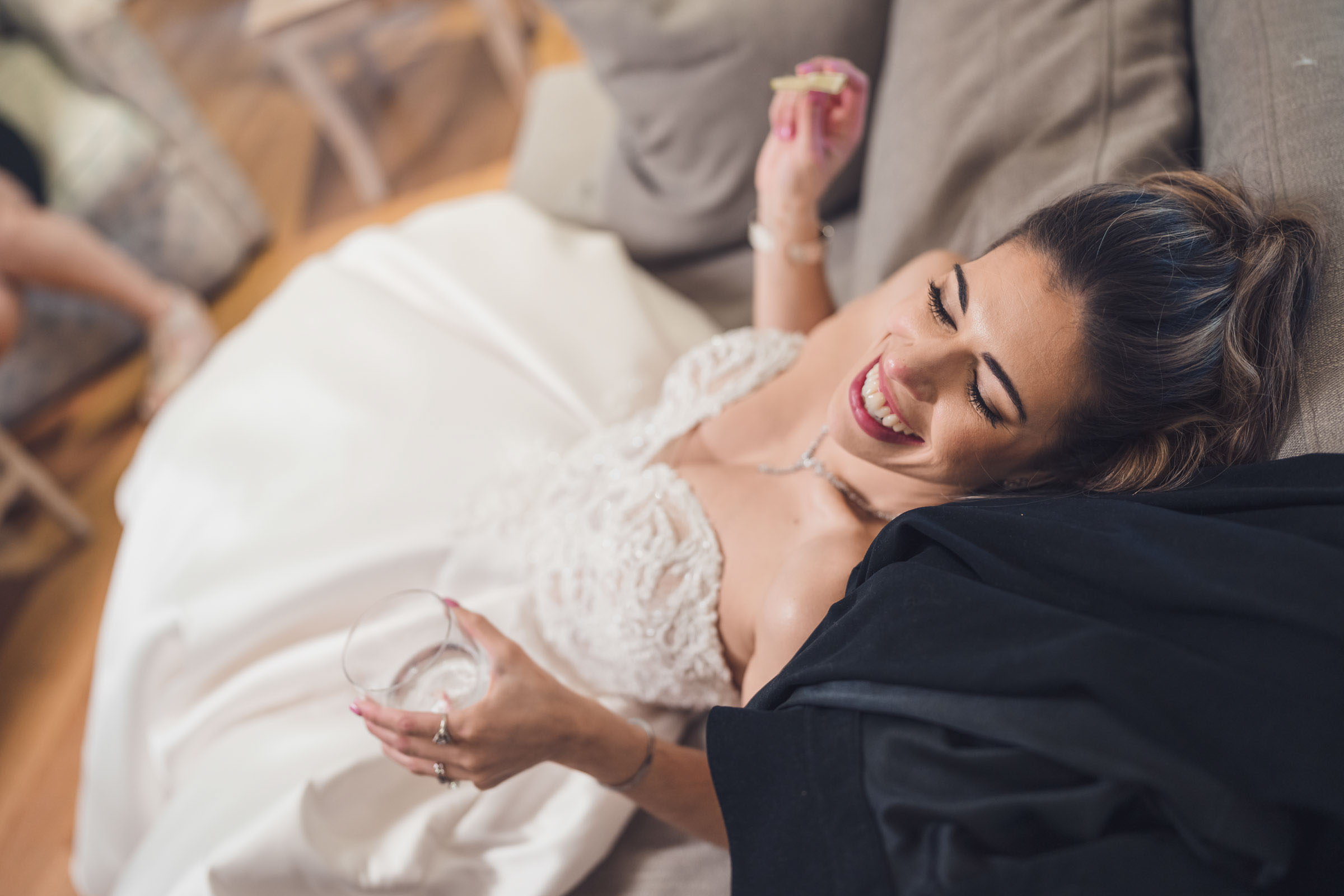 Bride relaxing on couch in wedding dress holding drink and smiling during reception at Alhambra Hall