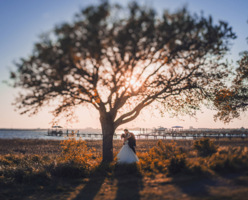 Bride and groom embracing under large oak tree at sunset with waterfront and dock in background at Alhambra Hall Mount Pleasant Charleston