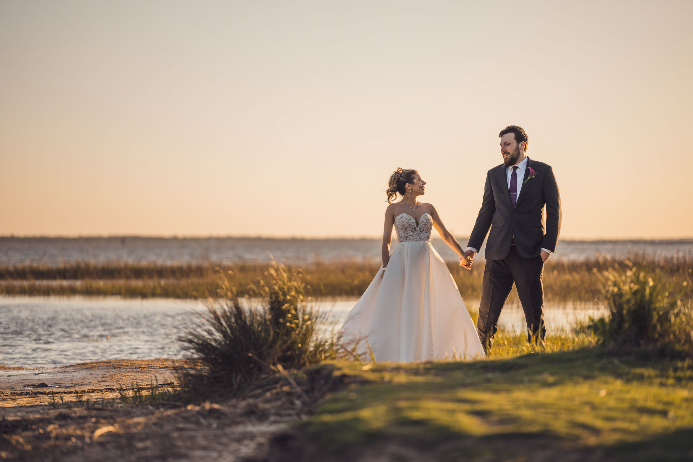 Bride and groom holding hands and walking along marsh waterfront at golden hour near Alhambra Hall Charleston wedding venue