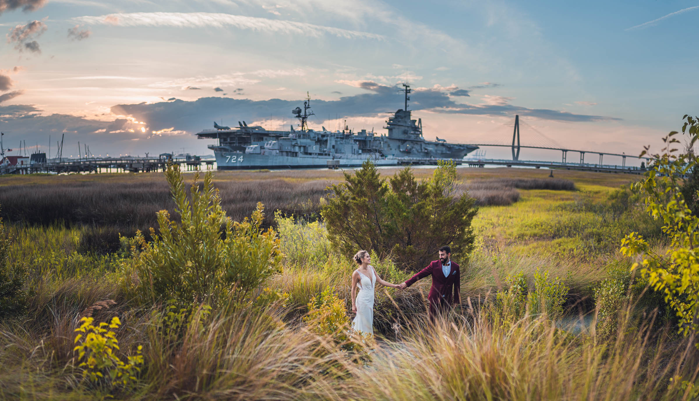 Newlyweds walk through marsh grasses with the USS York Town and Ravenel Bridge behind them at Charleston Harbor Resort.