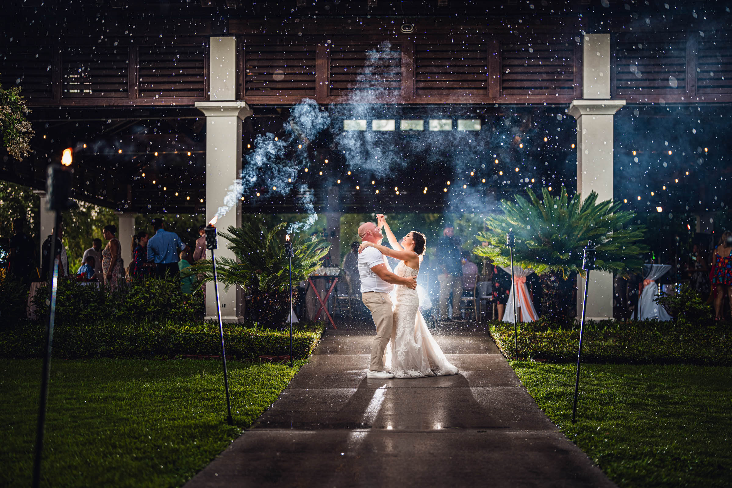 Newlyweds dance outside the Lookout Pavilion at night as rain falls around them beneath string lights.