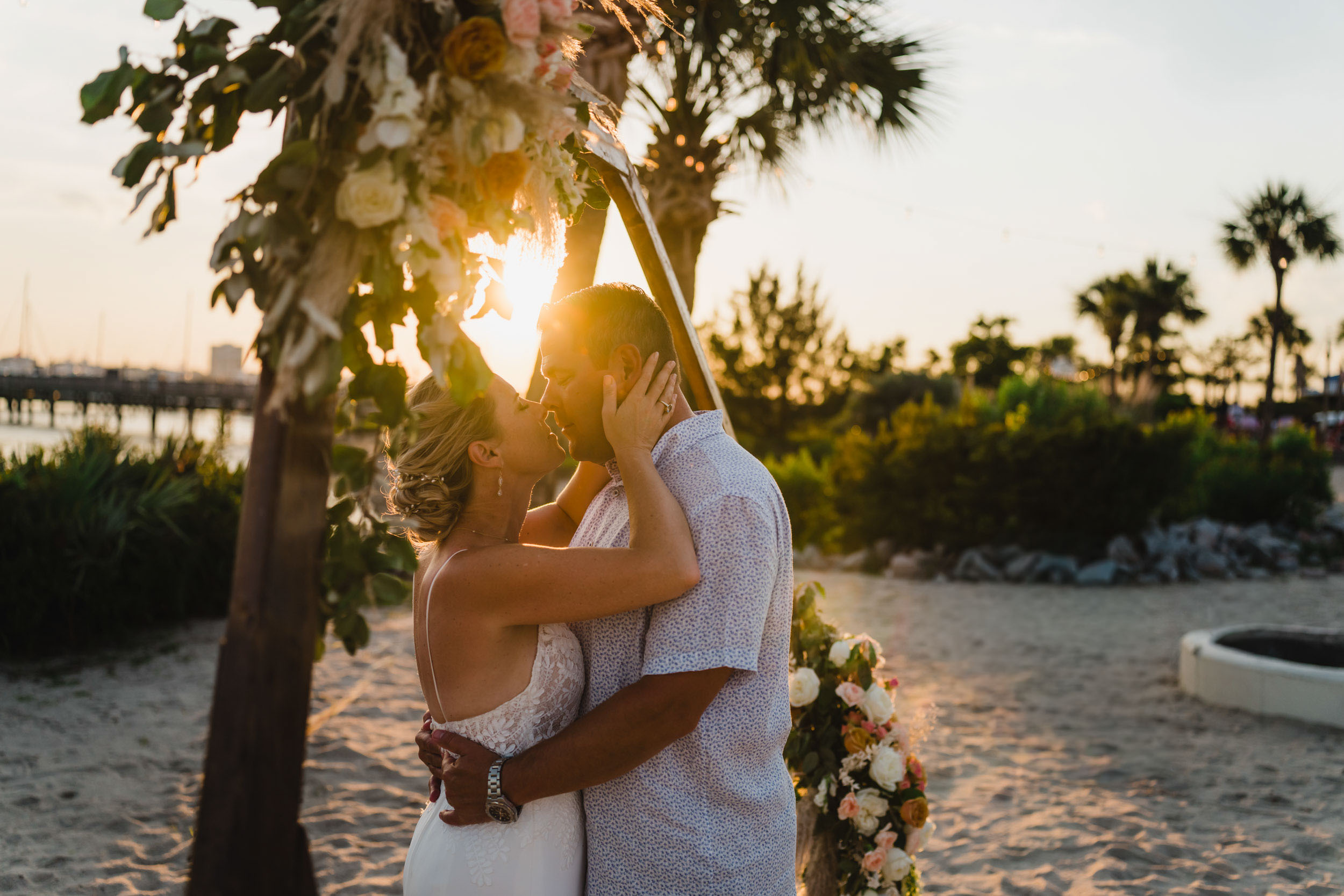 Bride and groom kiss beneath a flower-covered palm arch on the sandy waterfront at sunset.