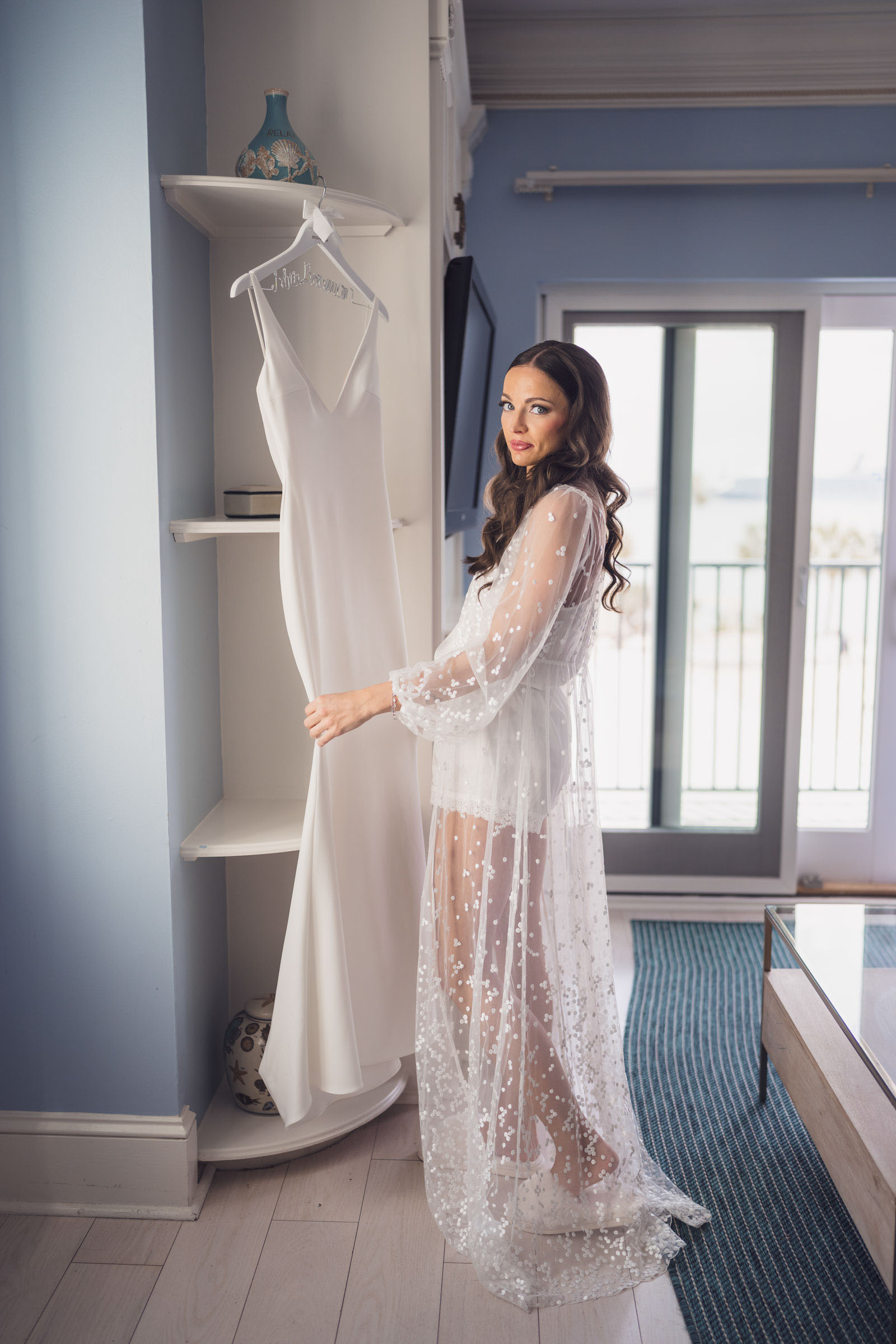 Bride in a sheer beaded getting-ready robe stands beside her wedding dress in a waterfront hotel room.