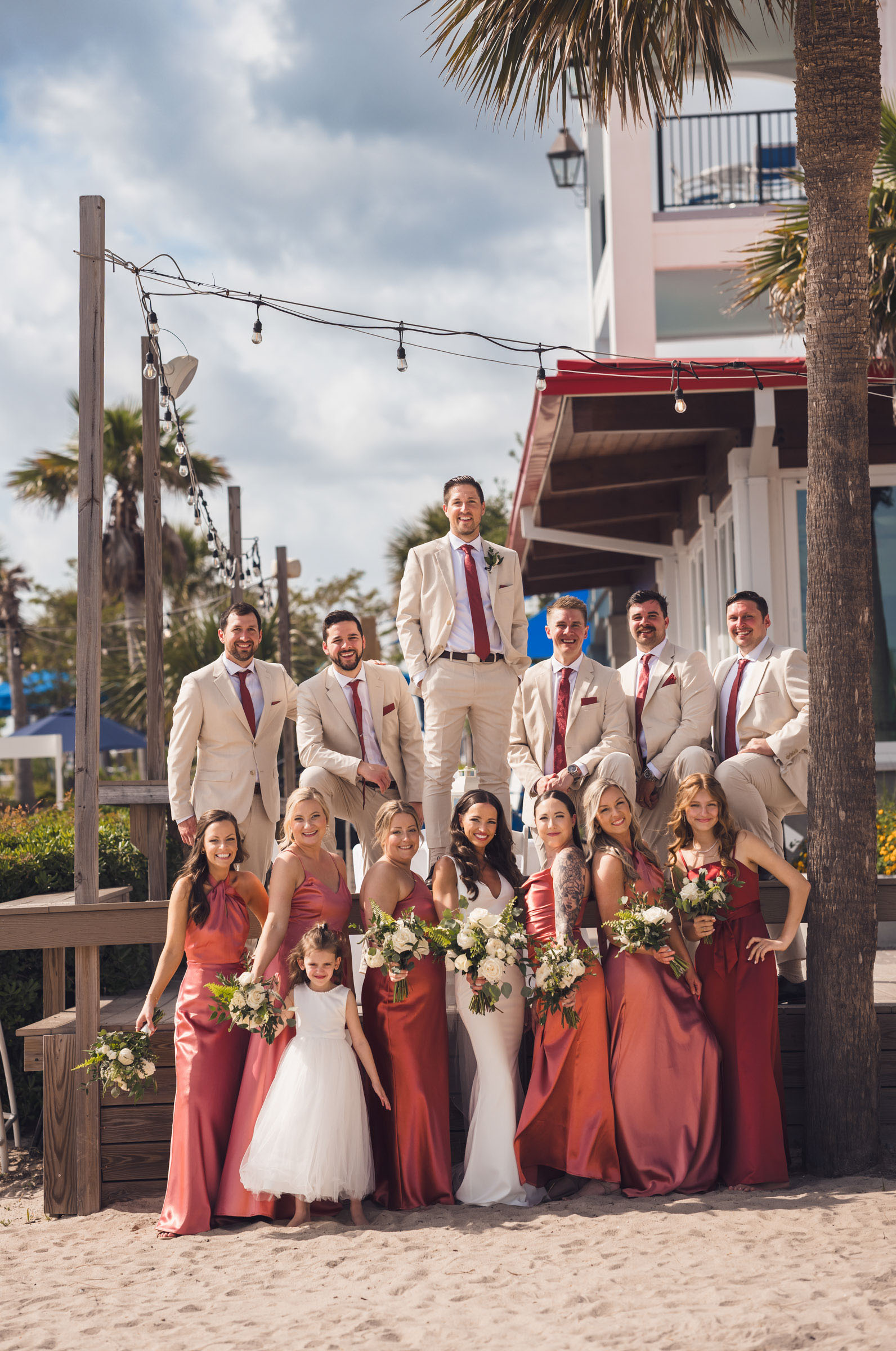 Bride, groom and wedding party pose together outside Charleston Harbor Resort & Marina with palm trees and string lights behind them.