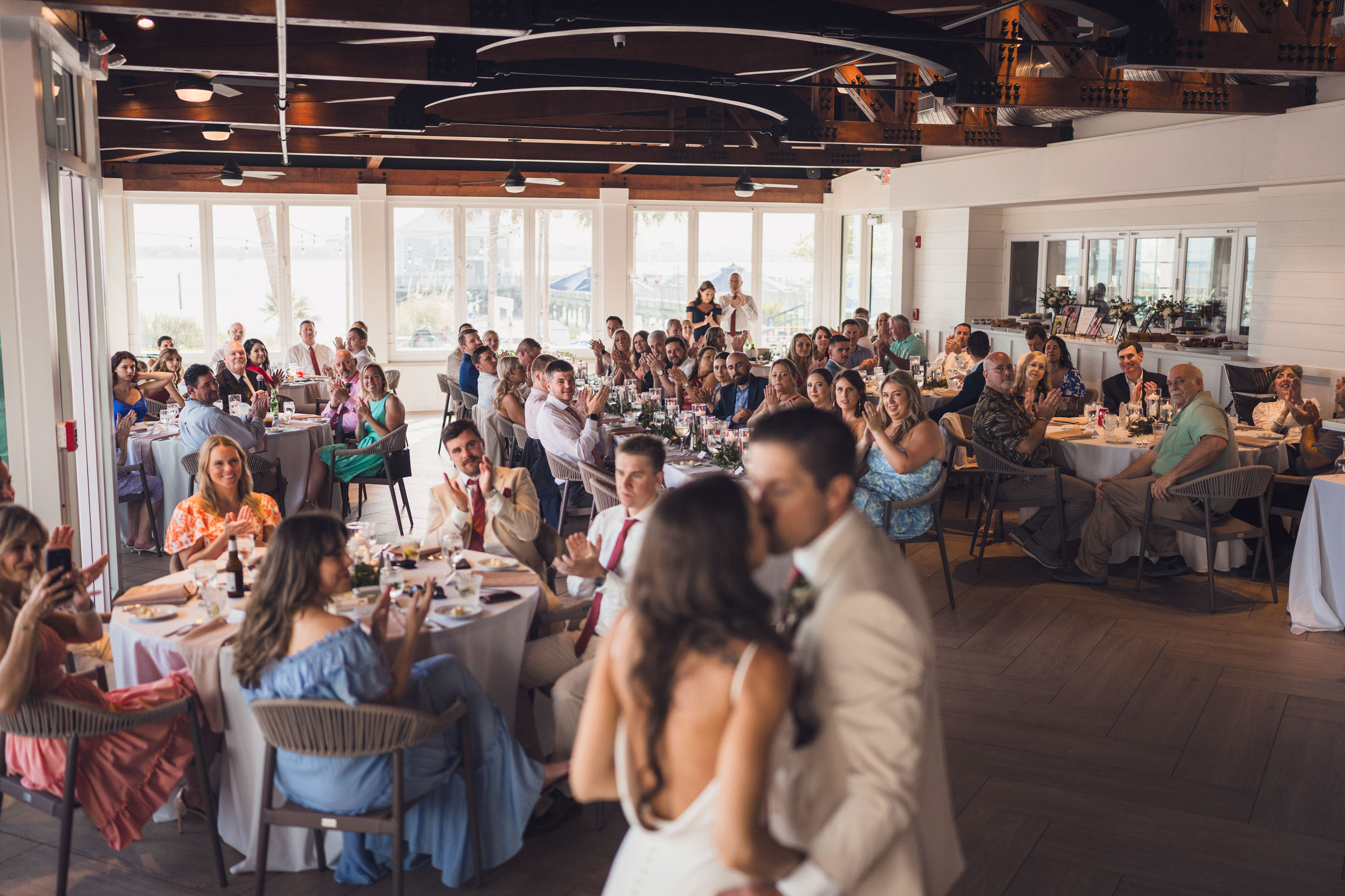 Newlyweds kiss in the foreground while wedding guests applaud inside the waterfront reception room.