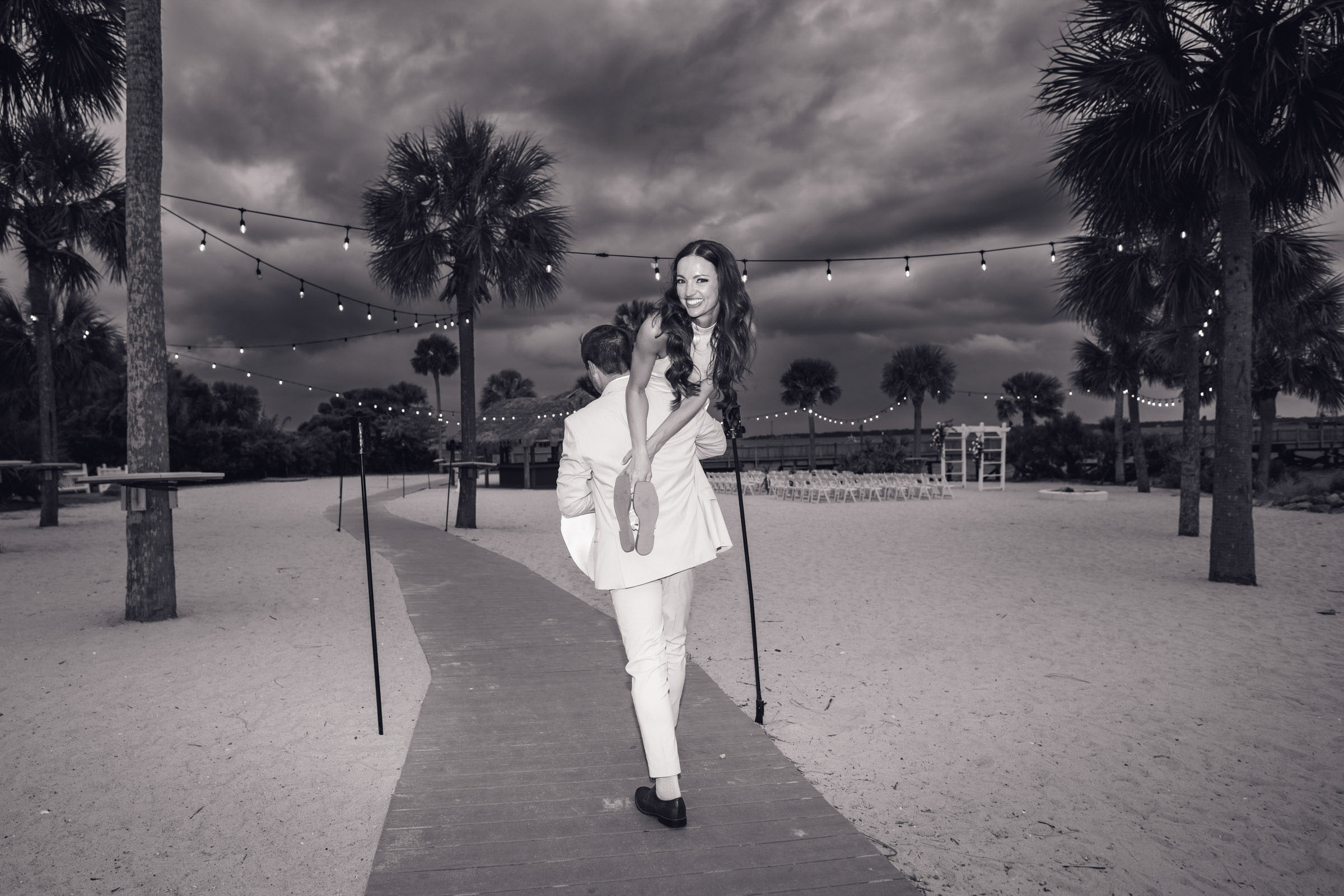Black-and-white photo of the groom carrying the bride piggyback along the sandy boardwalk beneath string lights and dark clouds.
