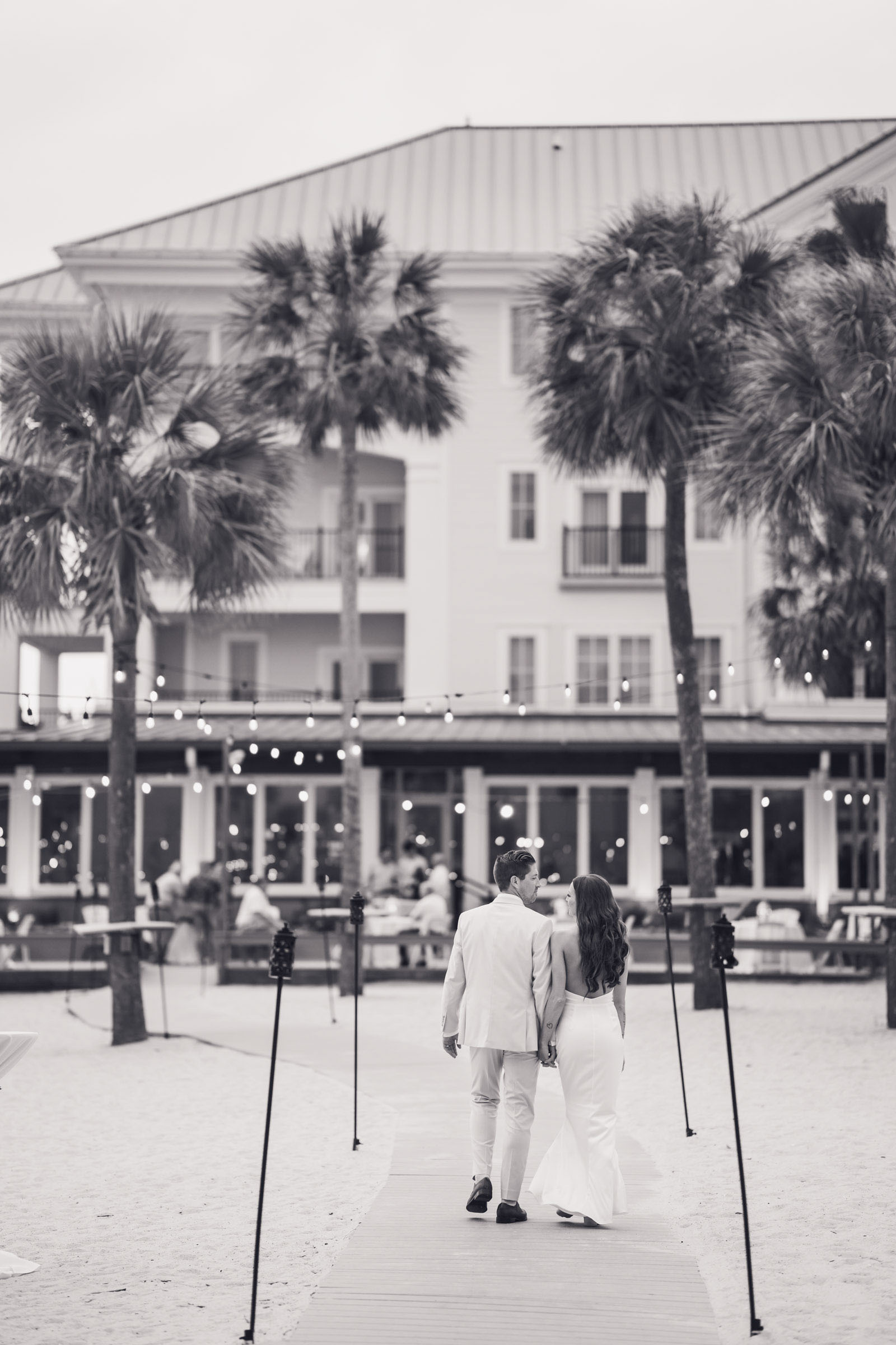 Black-and-white photo of newlyweds walking hand in hand along the boardwalk toward Charleston Harbor Resort & Marina.