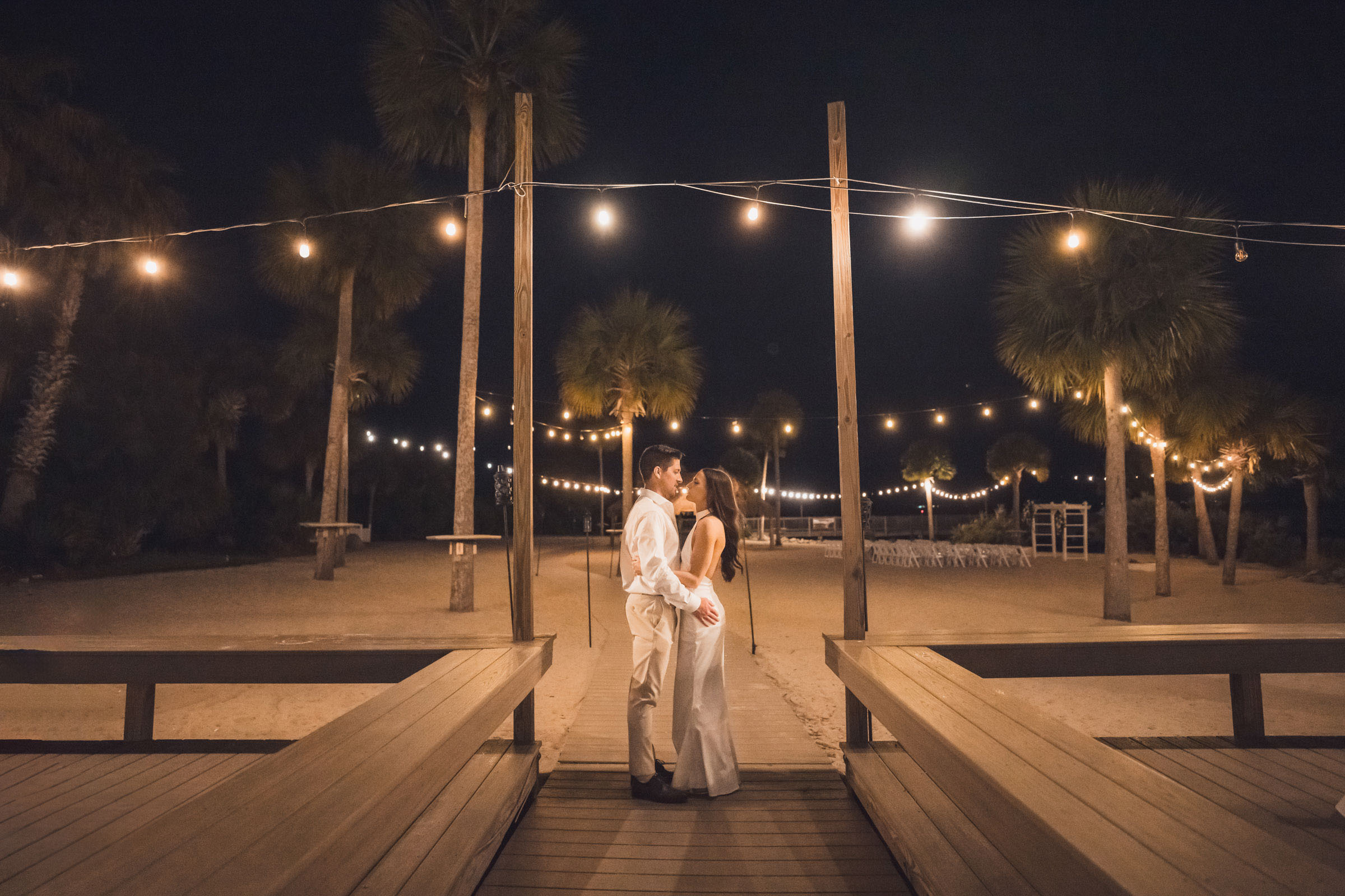 Bride and groom kiss beneath cafe lights on the waterfront boardwalk at night.