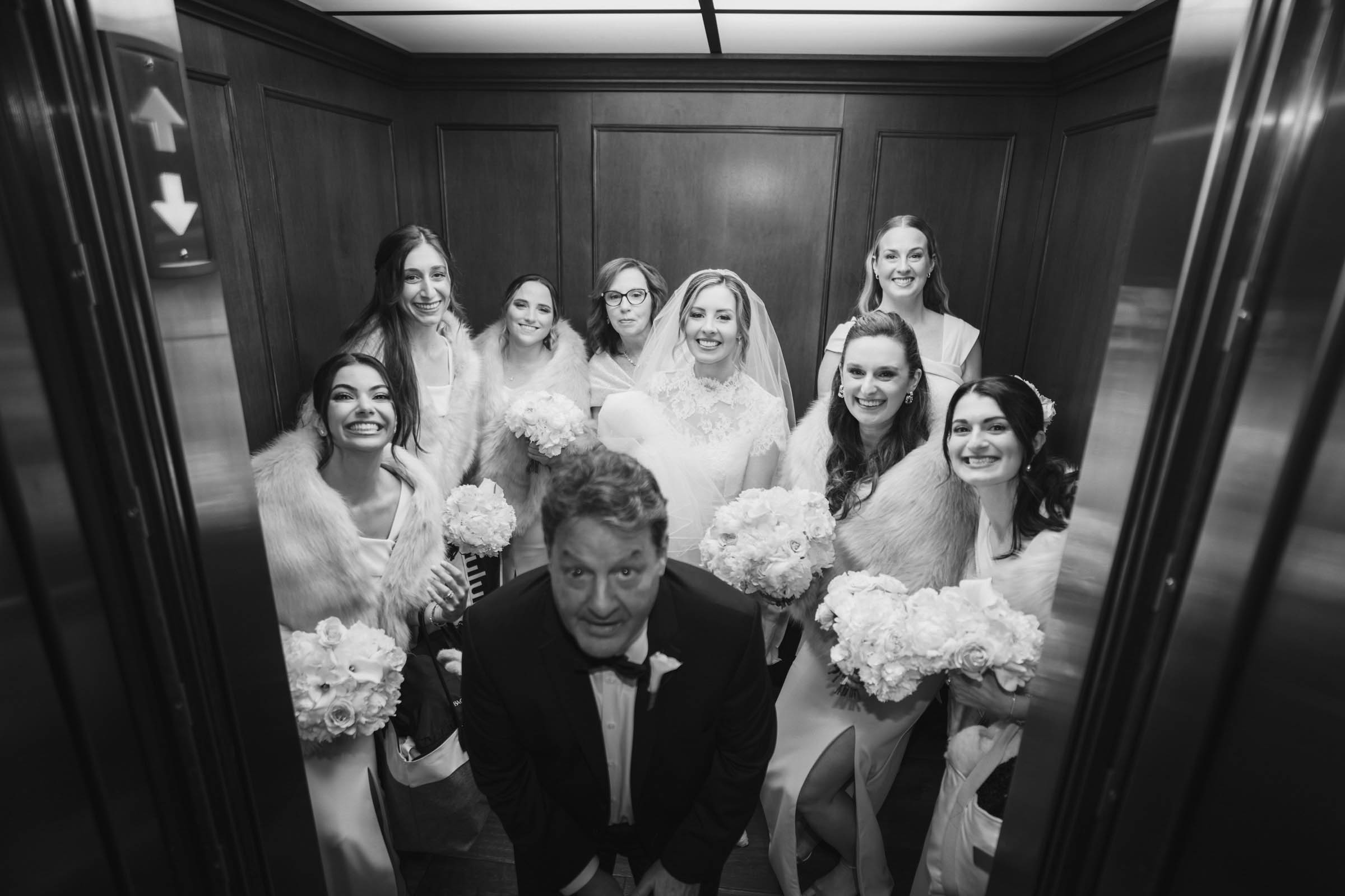 Black-and-white photograph of the bride, bridesmaids and family crowded together inside an elevator before the wedding.