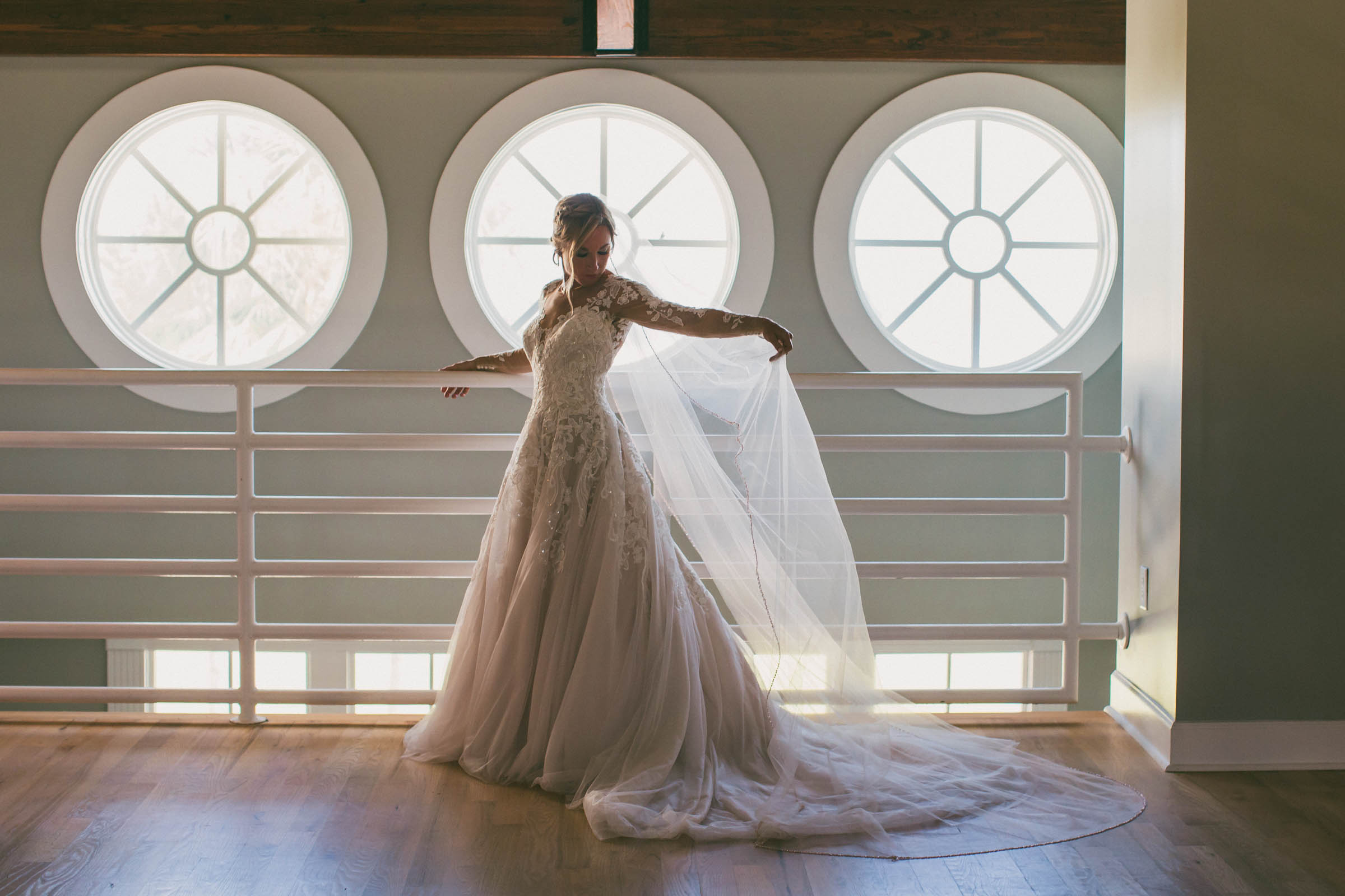 Bride in a lace wedding gown standing indoors at Citadel Beach Club in front of three large round windows, holding out her veil.