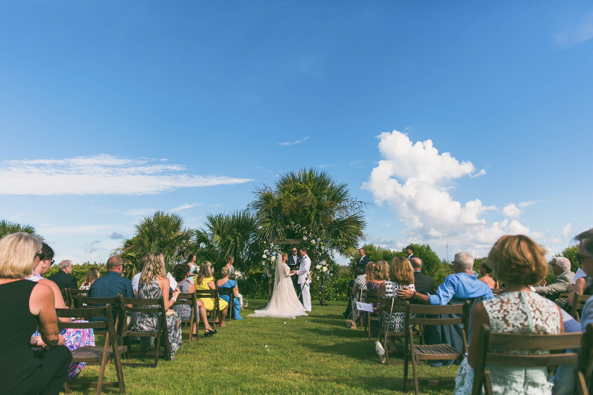 Guests seated on either side of a grassy aisle during a Citadel Beach Club wedding ceremony with palm trees and blue sky overhead.