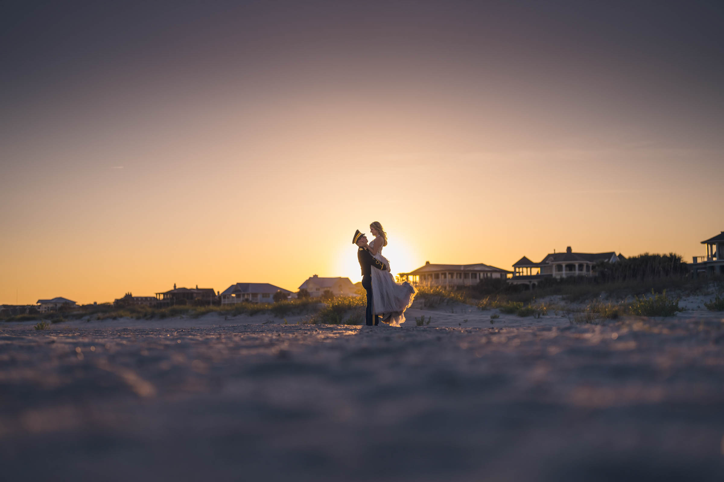 Silhouetted bride and groom embracing on the beach at sunset near Citadel Beach Club with houses visible in the distance.
