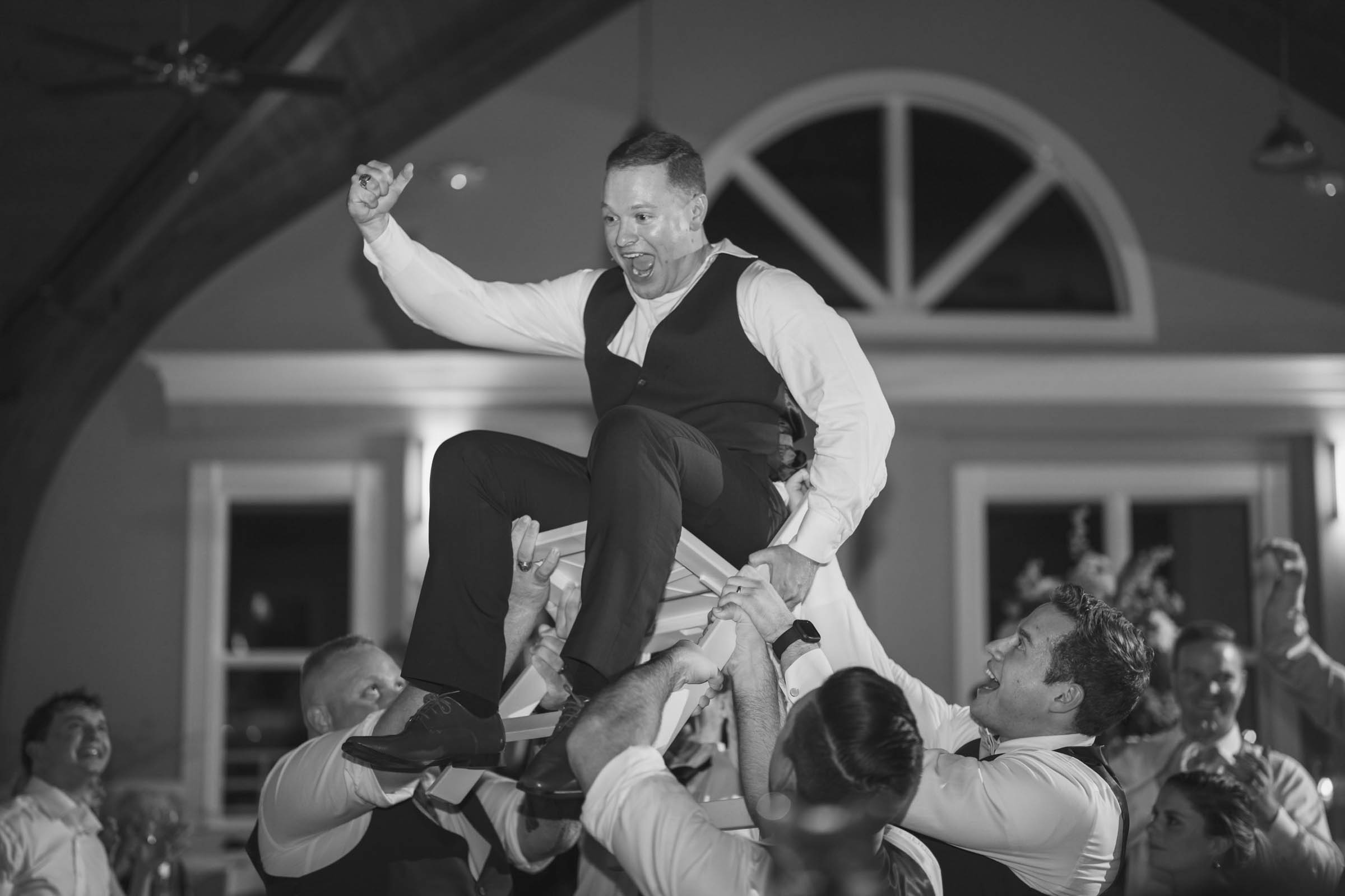 Black and white image of a groom being lifted in a chair by wedding guests during the reception at Citadel Beach Club.