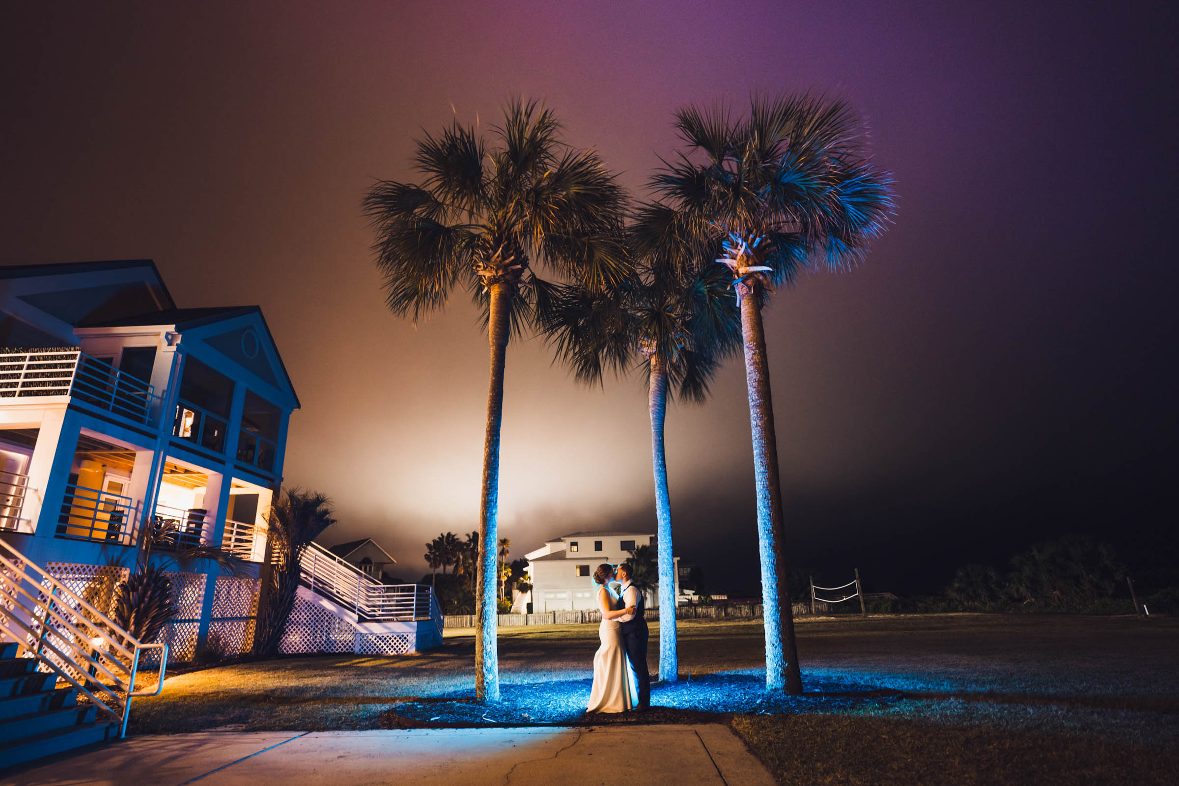 And after dark photo of a bride and groom standing under towering palm trees. They are lit in a magical blue light whit a warm orange and magenta sky above.