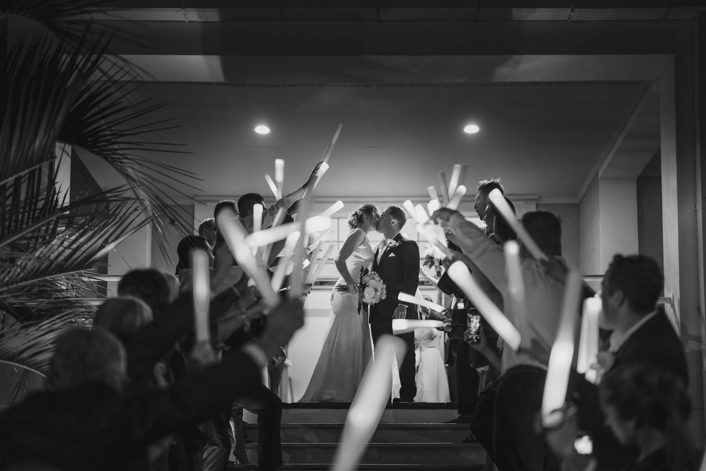 white photo of a bride and groom kissing beneath a tunnel of guests holding glow sticks outside Citadel Beach Club.