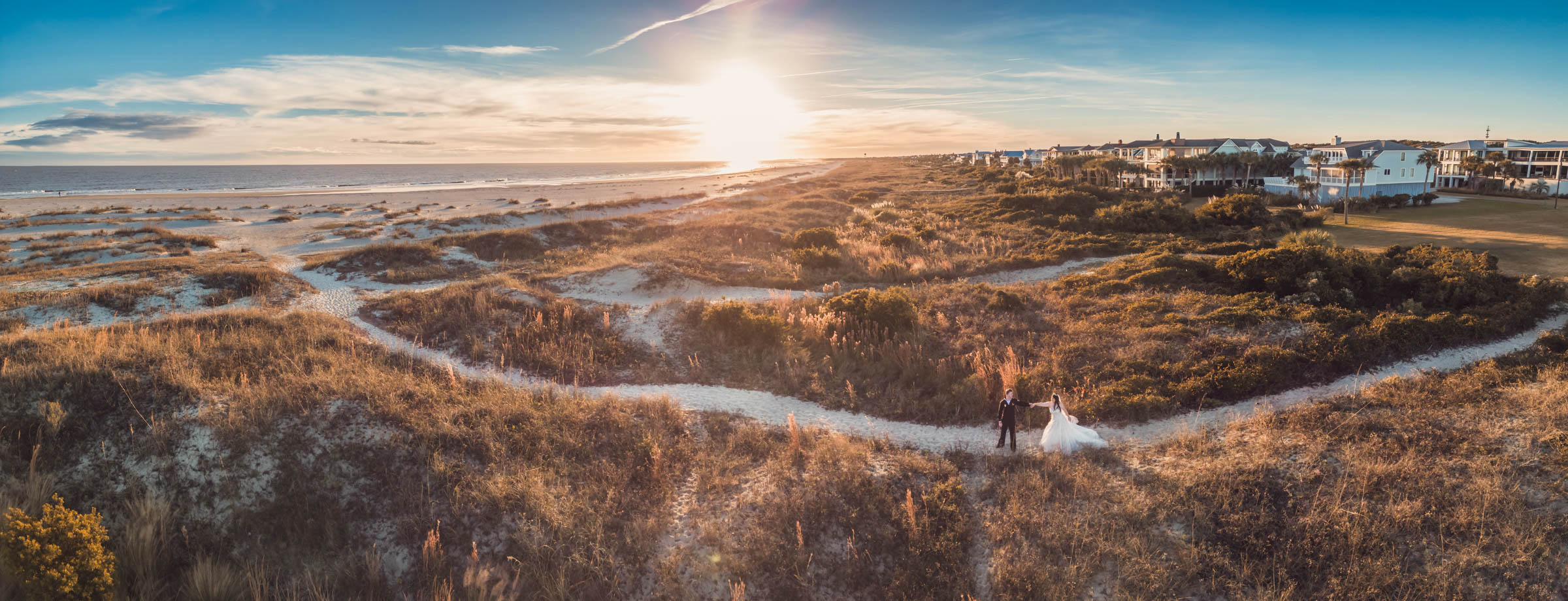 Aerial view of bride and groom standing on sandy paths through the dunes beside the beach at Citadel Beach Club during sunset.