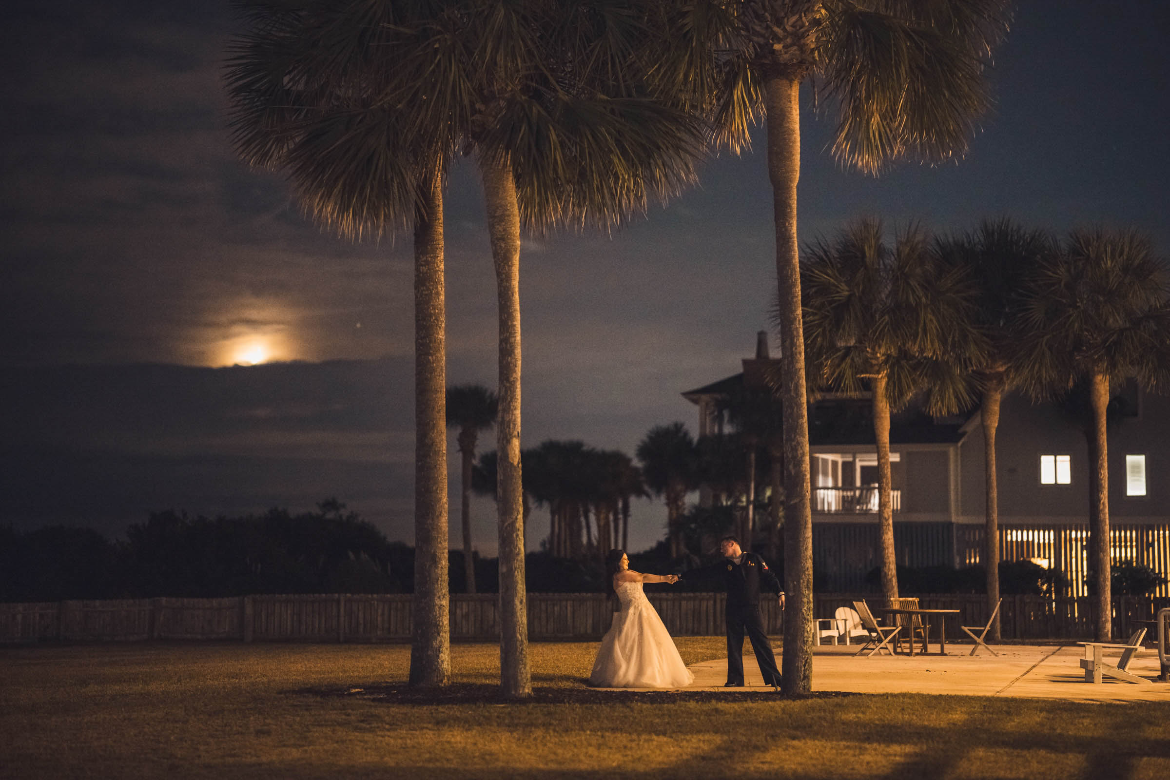 Bride and groom dancing outdoors at night near palm trees outside Citadel Beach Club with the moon rising in the background.