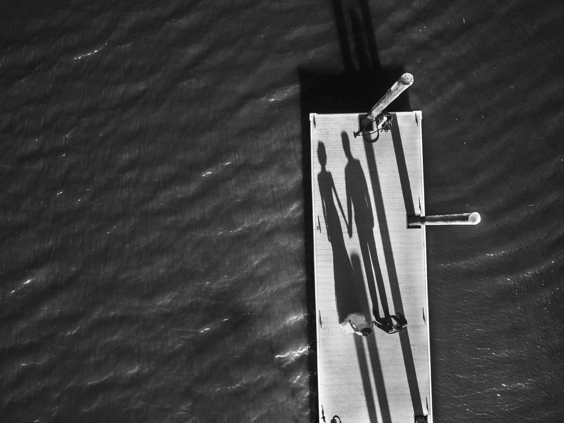 Birds Eye View Black-and-white aerial photograph of a bride and groom holding hands on the dock at Creek Club at I’On, their long shadows stretching across the boards and marsh water.