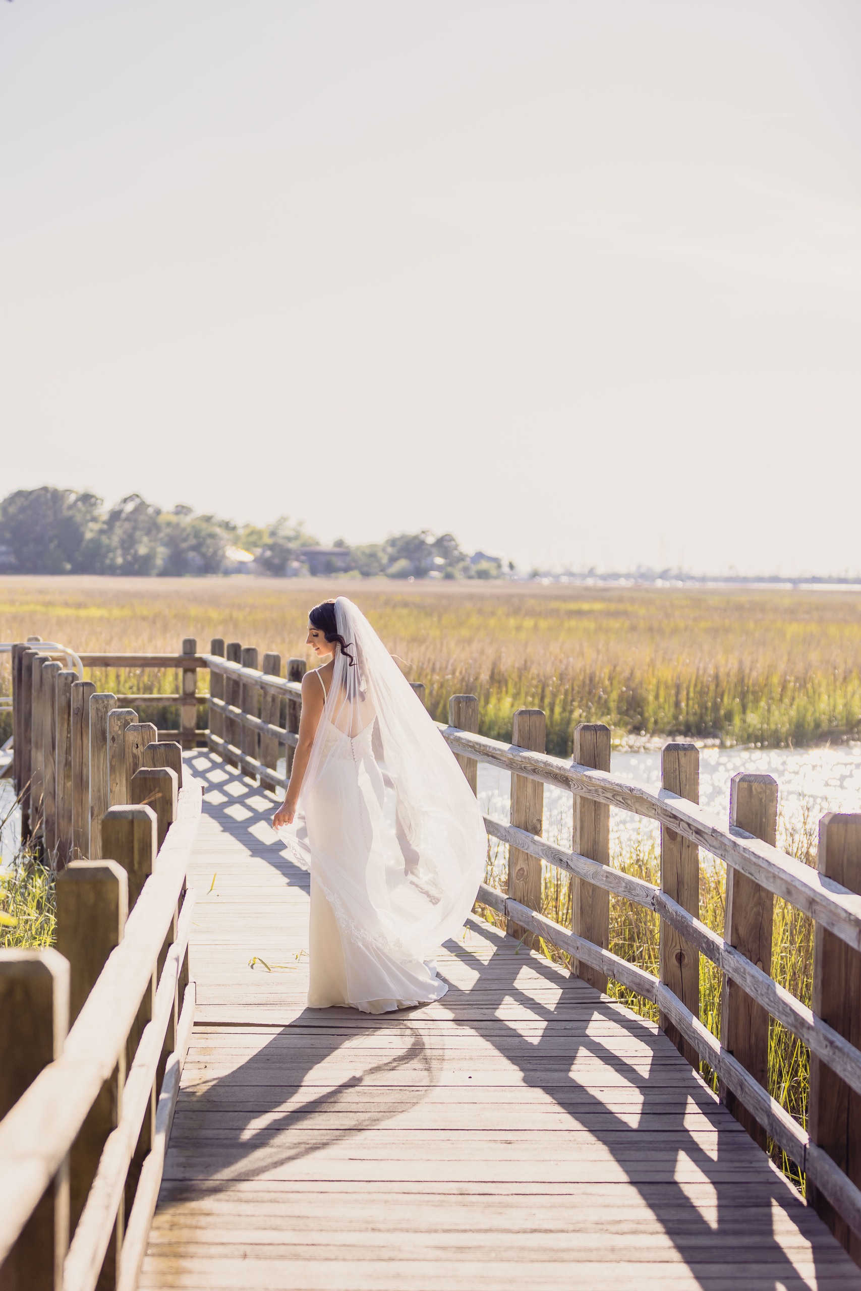 angel Bride walking along the wooden boardwalk through the marsh at Creek Club at I’On, wearing a long veil in bright afternoon light.