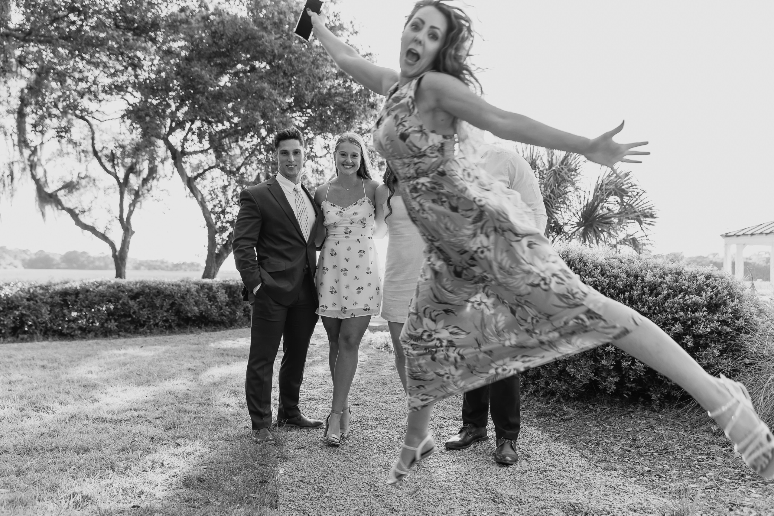 Formal Portrait, Briefly Black-and-white candid of a wedding guest jumping into the foreground during group portraits outside Creek Club at I’On.