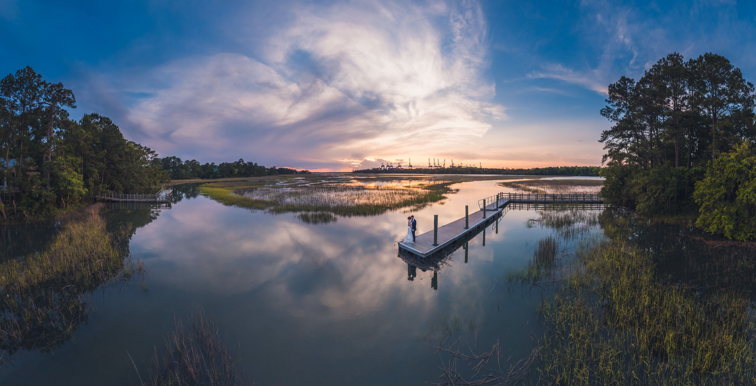 worth a little walk Aerial sunset portrait of a bride and groom standing on the dock at Creek Club at I’On surrounded by reflective marsh water in Mount Pleasant.
