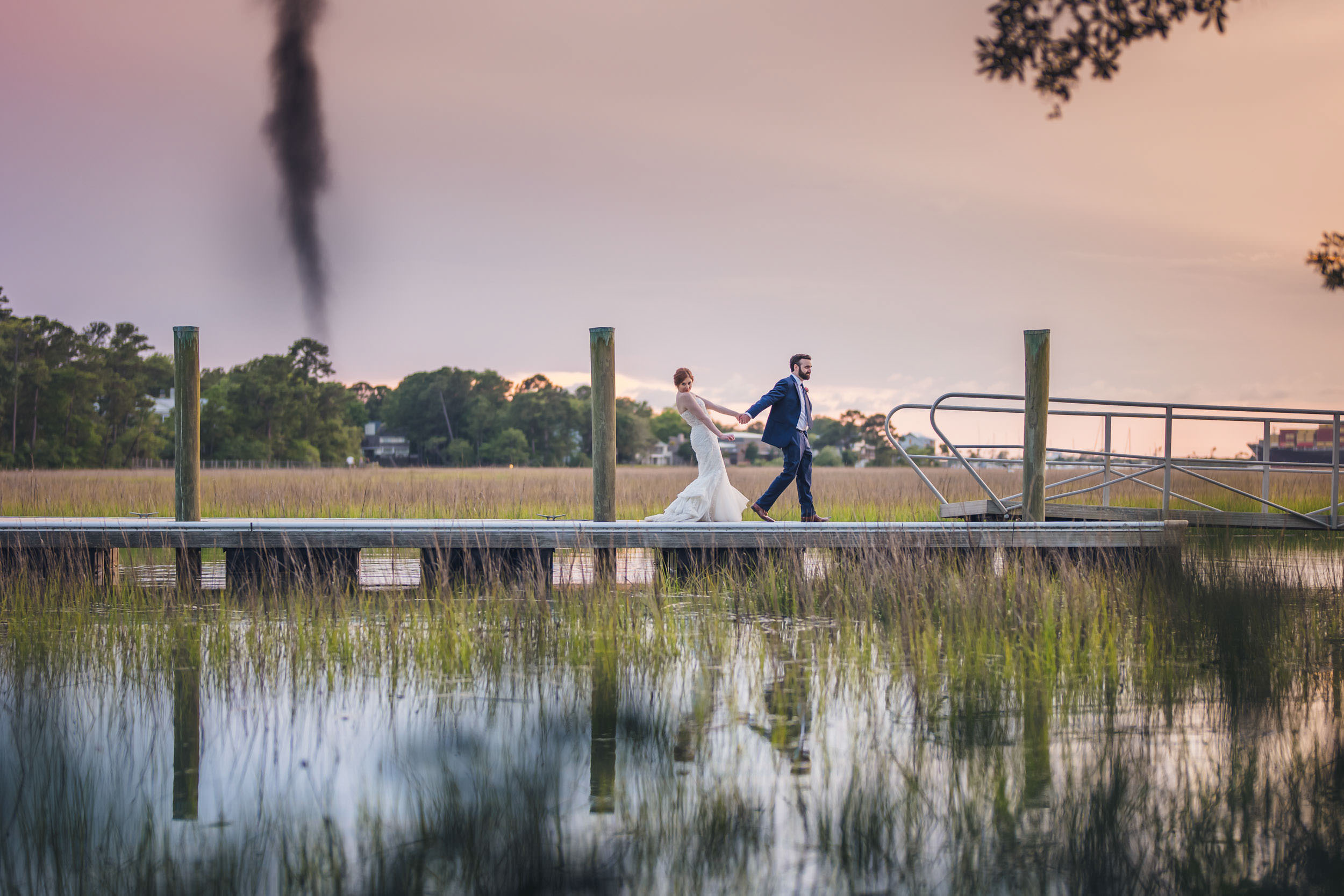 be yourself A groom leading his bride along the dock at Creek Club at I’On with marsh grass and water surrounding them at dusk. She is looking playfully at the camera.