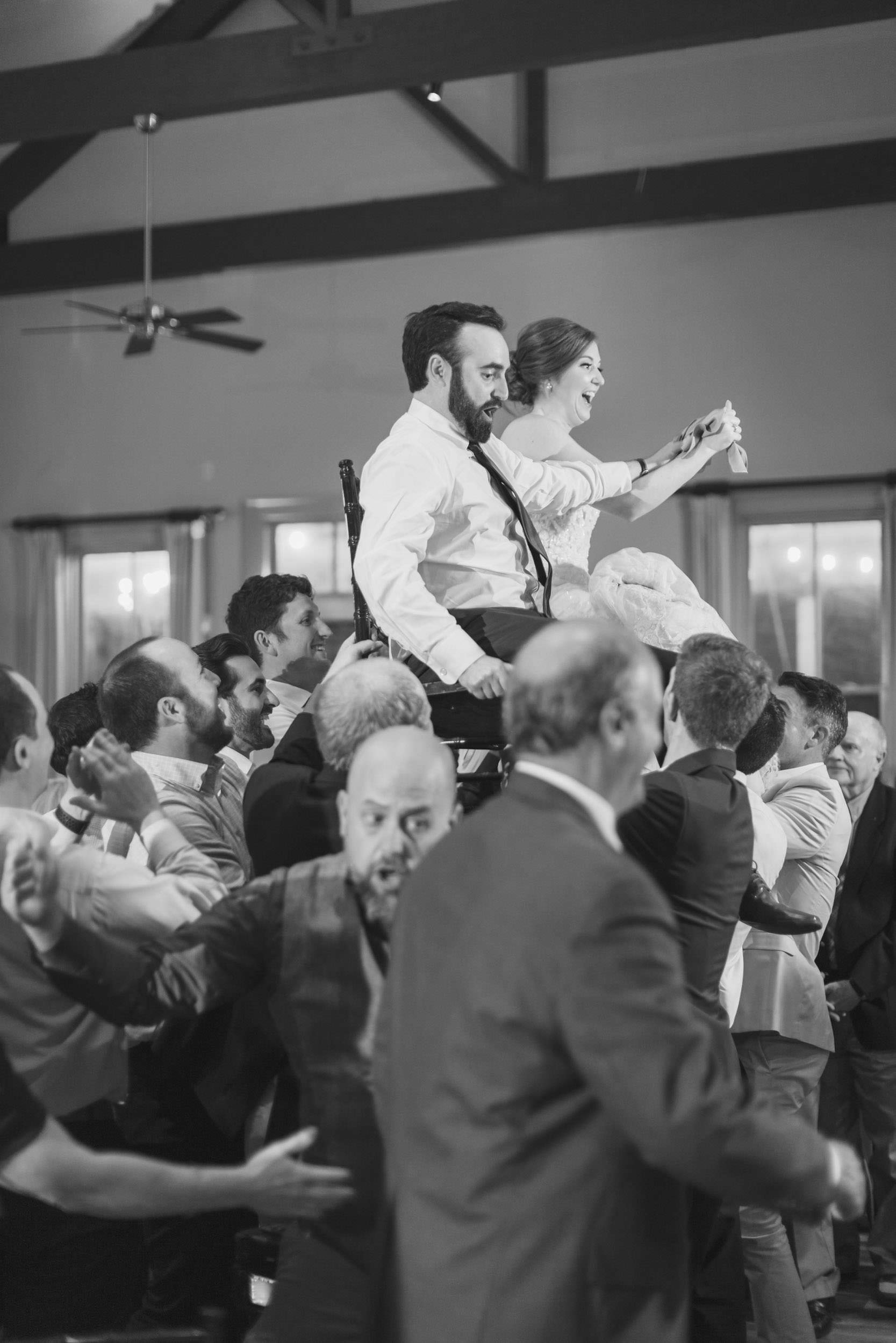 Up We Go Black-and-white photograph of a bride and groom lifted in chairs above their wedding guests during a lively reception at Creek Club at I’On.