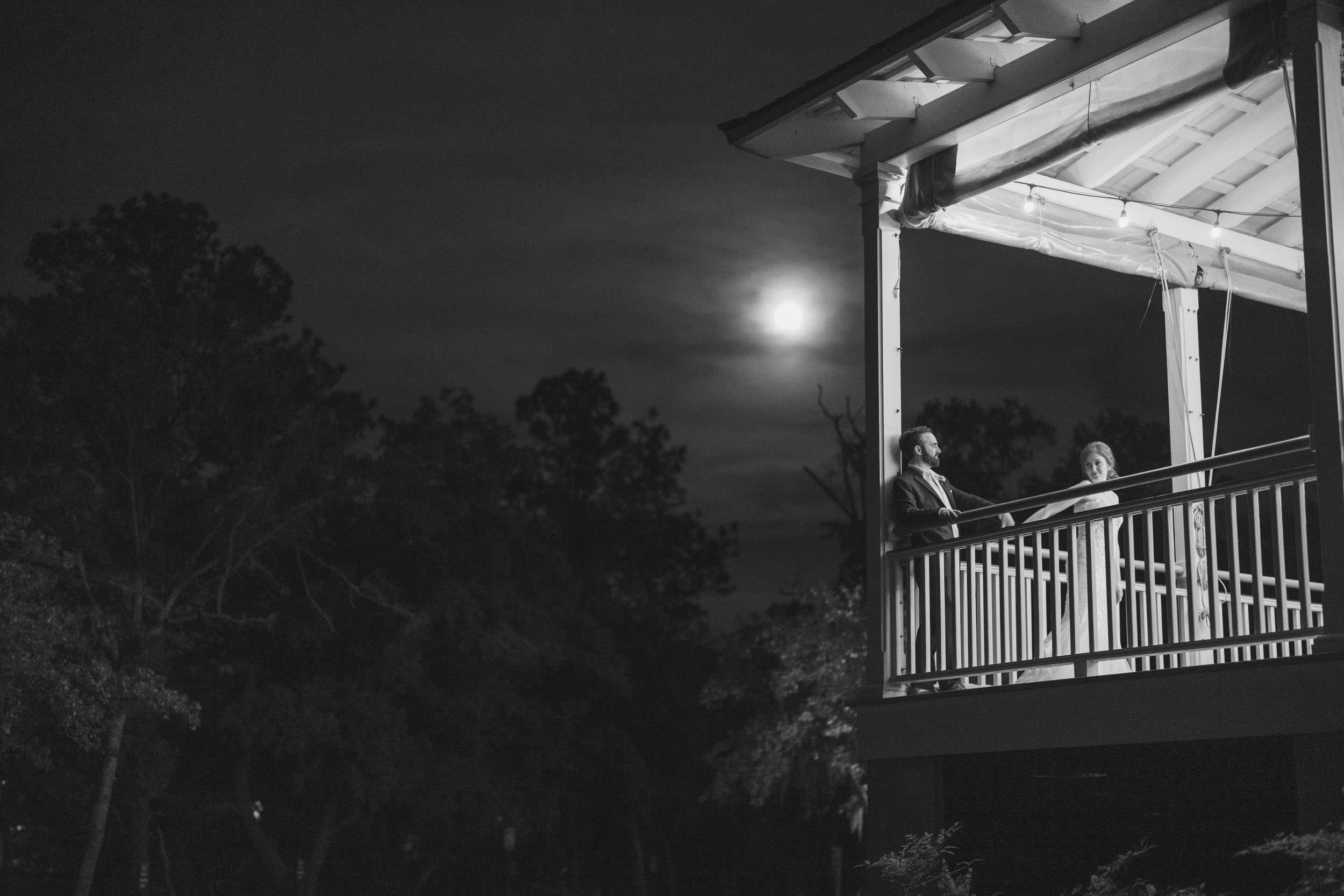 Moon Break Black-and-white nighttime portrait of a bride and groom standing on the covered porch at Creek Club at I’On beneath a bright moon.