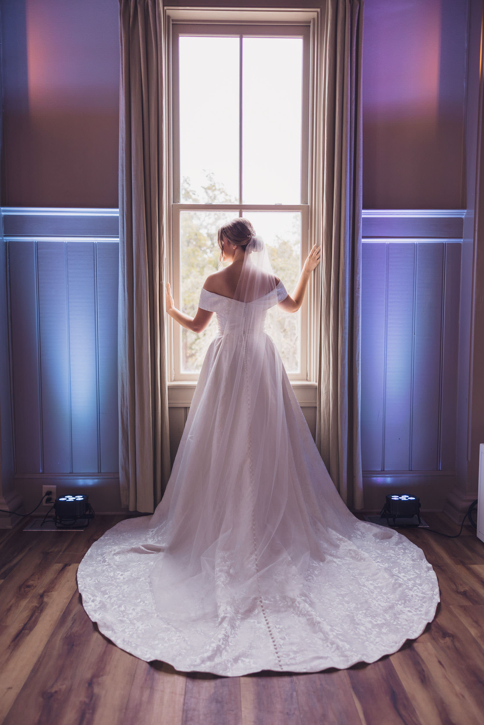 Windows and Uplights Bride in her wedding gown and veil standing in front of a tall window inside Creek Club at I’On before the ceremony.