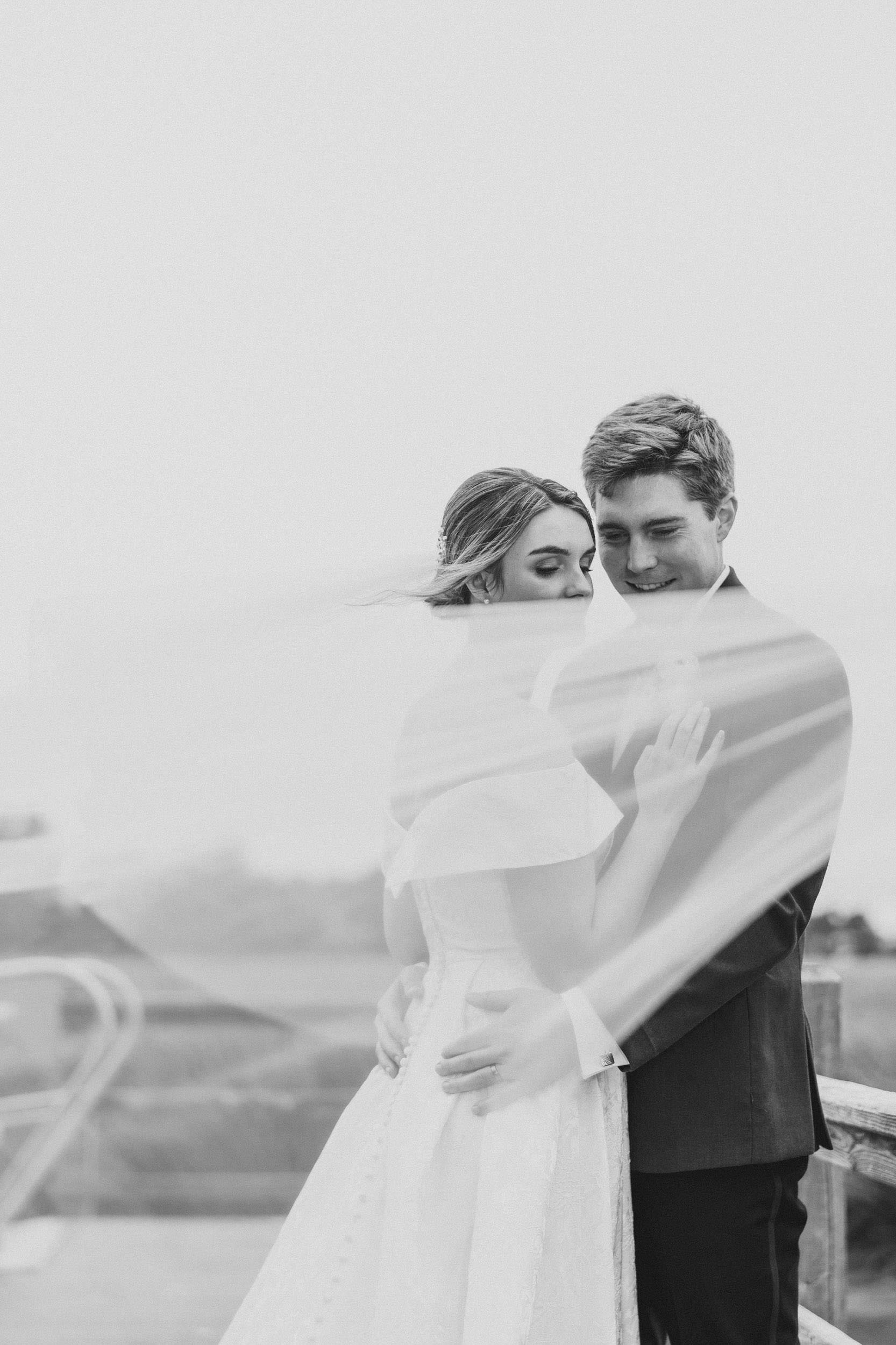 Windy Day Soft black-and-white portrait of a newlywed couple embracing as the bride’s veil blows in the wind near the marsh at Creek Club at I’On.