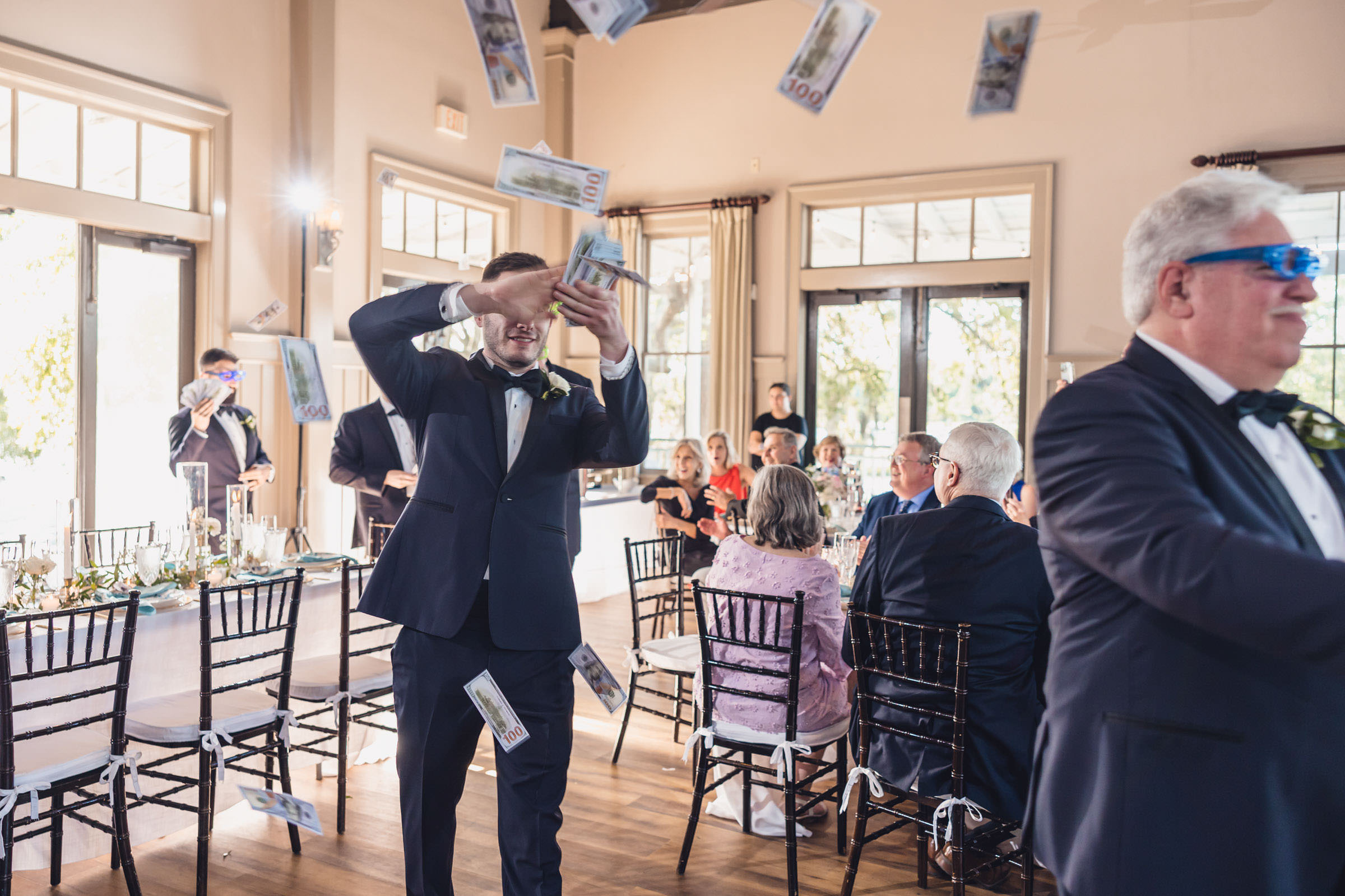 Making It Rain Wedding guest in a tuxedo tossing novelty bills into the air during a reception inside Creek Club at I’On.