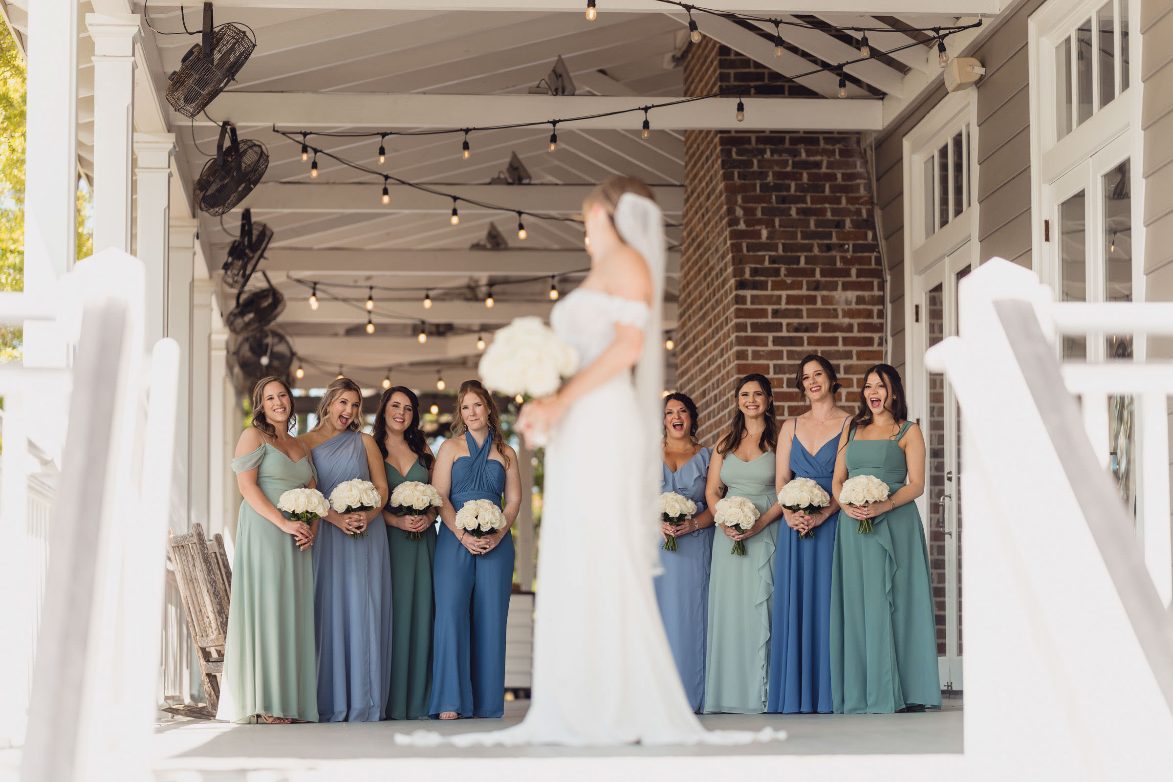 Audience Approved Bride standing on the covered porch at Creek Club at I’On as her bridesmaids smile and react to seeing her in her wedding dress.