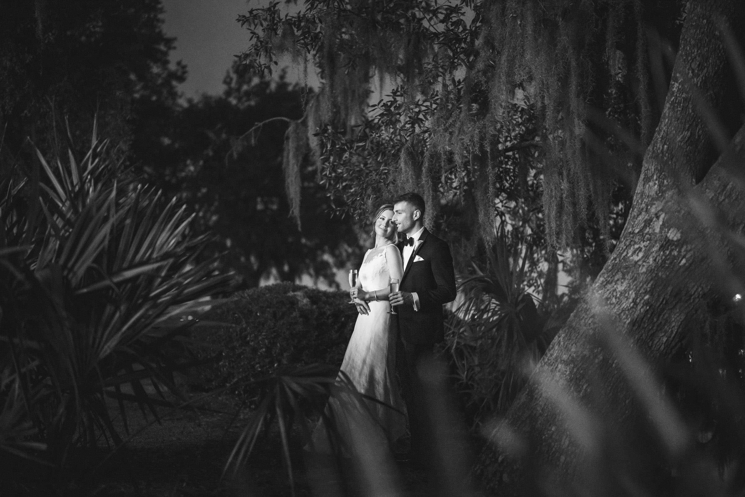 Champagne Break Black-and-white nighttime portrait of a bride and groom holding champagne beneath live oaks and Spanish moss at Creek Club at I’On.