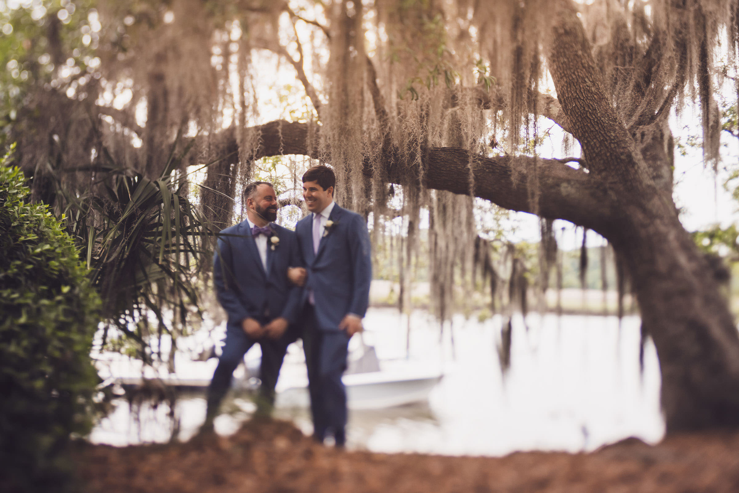 Mossy Beard Two grooms in blue suits standing together beneath live oaks and Spanish moss beside the marsh at Creek Club at I’On.