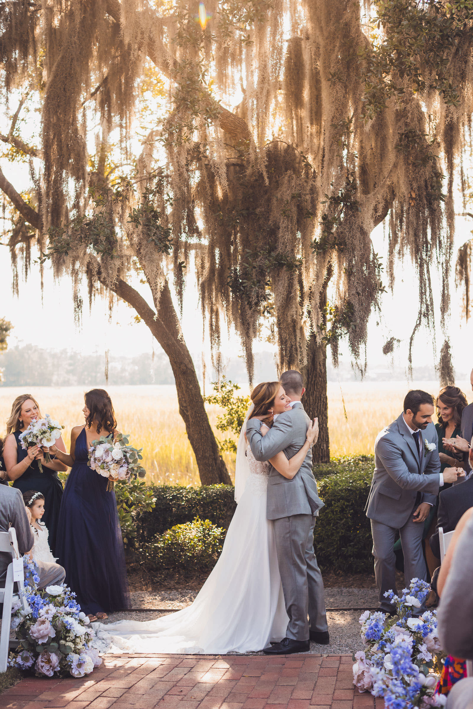 Oh the Glow Bride and groom embracing during an outdoor wedding ceremony beneath Spanish moss and live oaks at Creek Club at I’On.