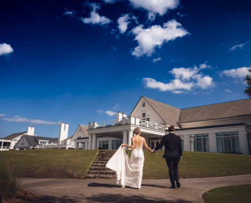 Newlyweds walking hand in hand toward the white clubhouse at Daniel Island Club beneath a bright blue Charleston sky.