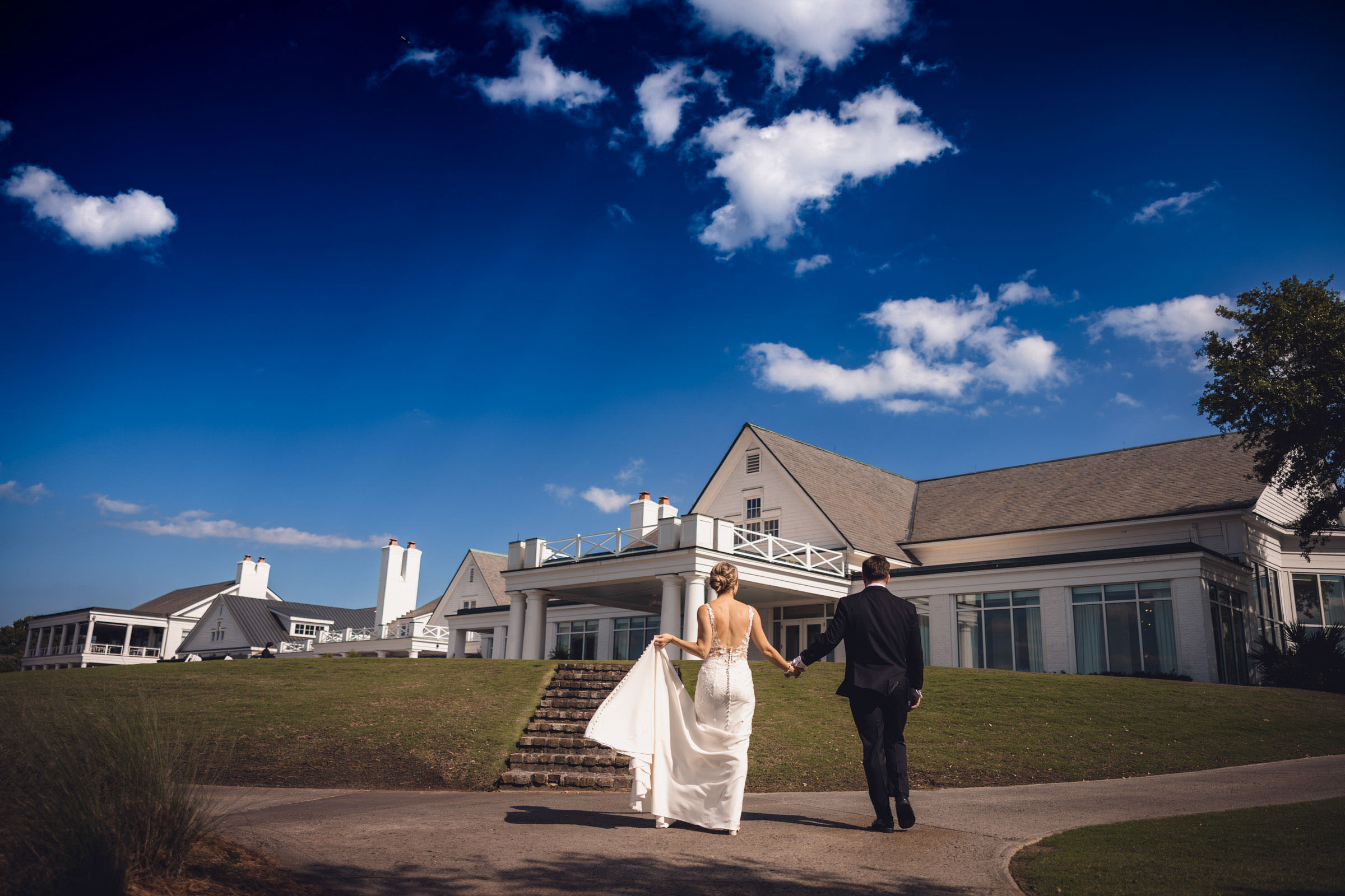 Daniel Island Club Wedding Newlyweds walking hand in hand toward the white clubhouse at Daniel Island Club beneath a bright blue Charleston sky.