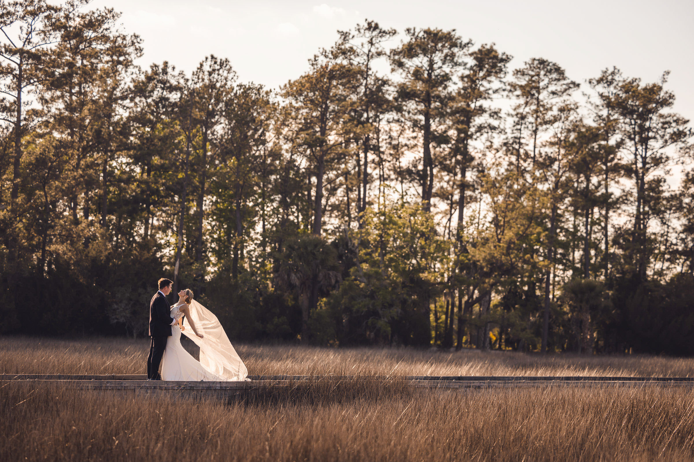 Windy Marsh Wedding Portrait Newlyweds on the Daniel Island Club boardwalk as the bride's veil catches the wind above the marsh grass.