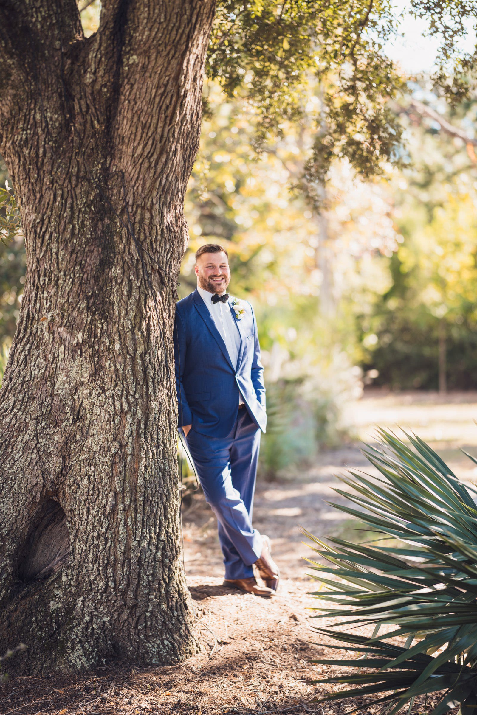 Groom Portrait at Daniel Island Club Groom in a blue suit leaning beside a large oak tree in the afternoon light at Daniel Island Club.
