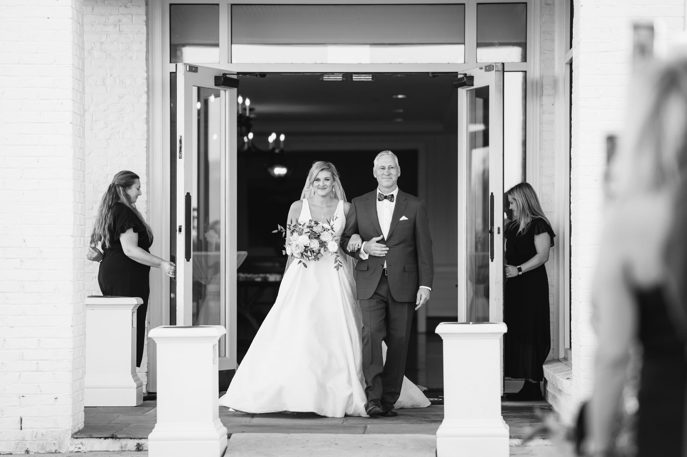 Bride and Father Enter the Ceremony Bride walking arm in arm with her father through the clubhouse doors toward an outdoor Daniel Island Club wedding ceremony.
