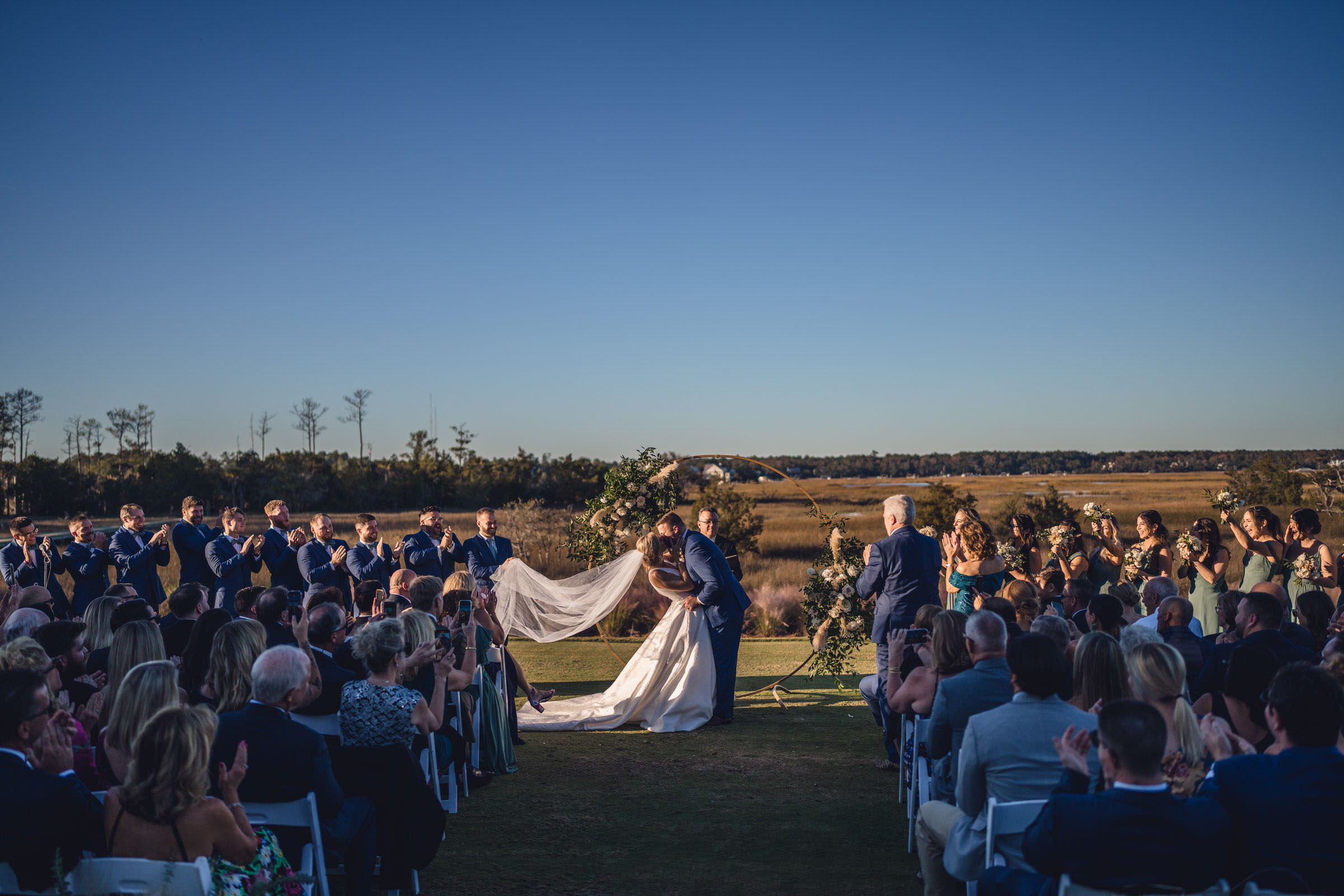 Just Married at Daniel Island Club Bride and groom kissing beneath their floral ceremony arch while wedding guests cheer beside the Daniel Island Club marsh.