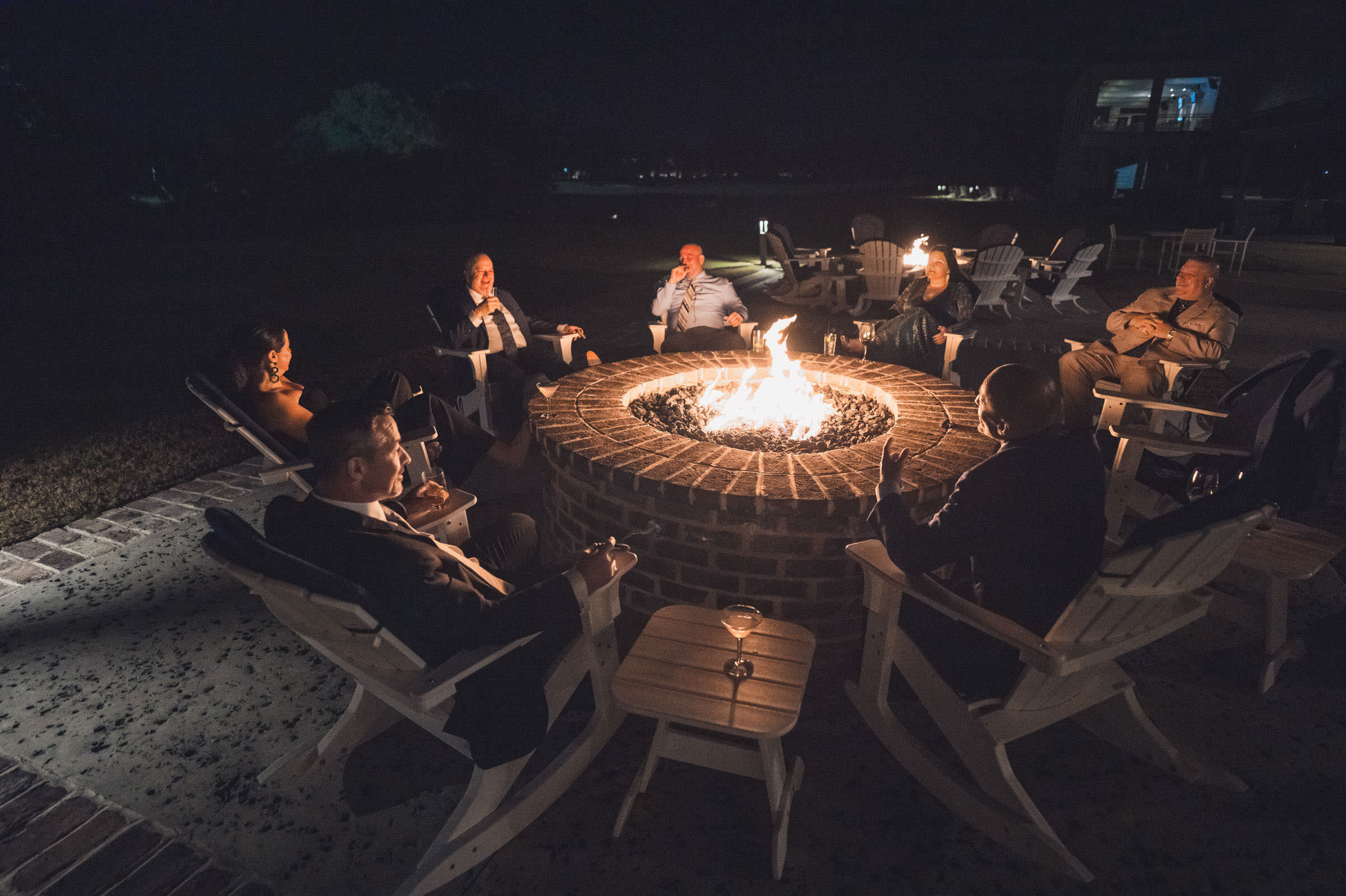 Fire Pit After the Wedding Wedding guests relaxing in Adirondack chairs around a large outdoor fire pit at Daniel Island Club after dark.