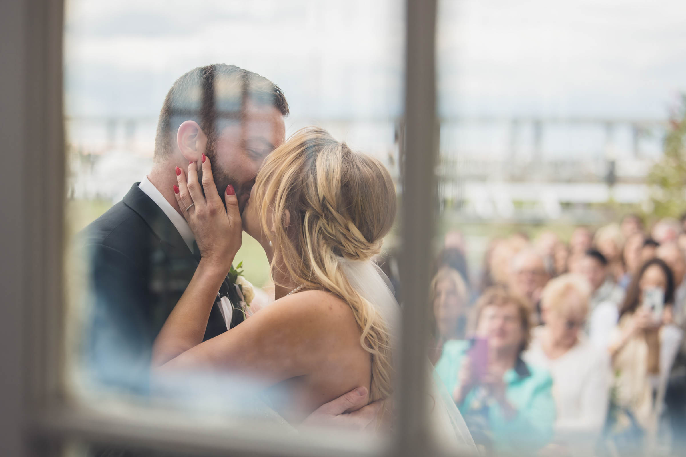 Historic Rice Mill First Kiss Newlyweds kiss after their ceremony, photographed through a window with their wedding guests gathered behind them.