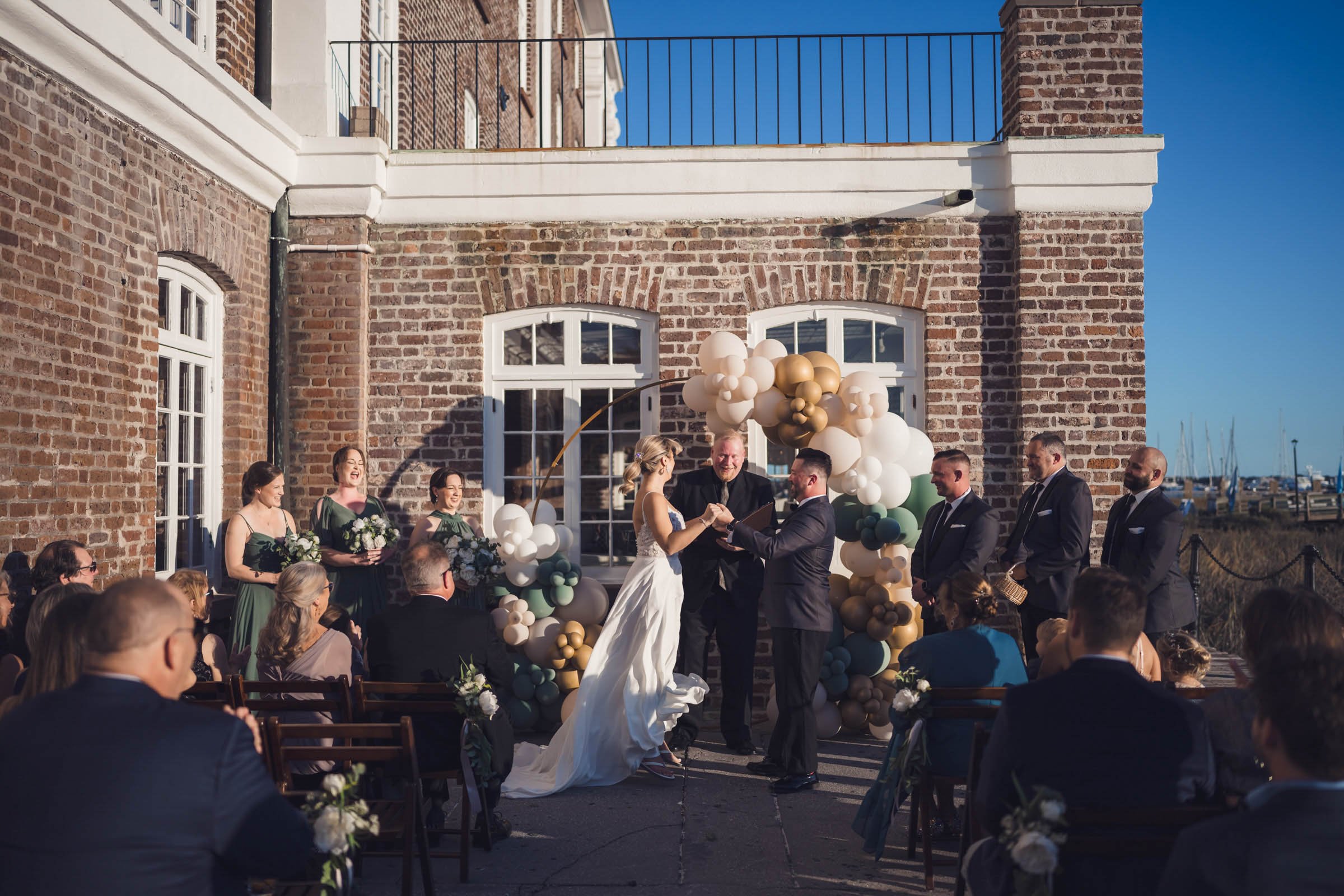 Outdoor Wedding Ceremony at the Historic Rice Mill Bride and groom exchange vows beside the brick Historic Rice Mill with wedding guests and the marina in the background.