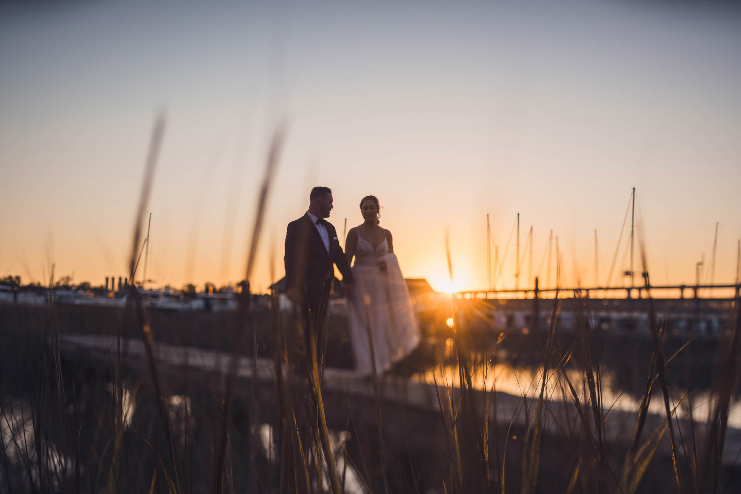 Historic Rice Mill Sunset Portrait Bride and groom walk through the marsh grass at sunset with sailboats and the Charleston marina behind them.